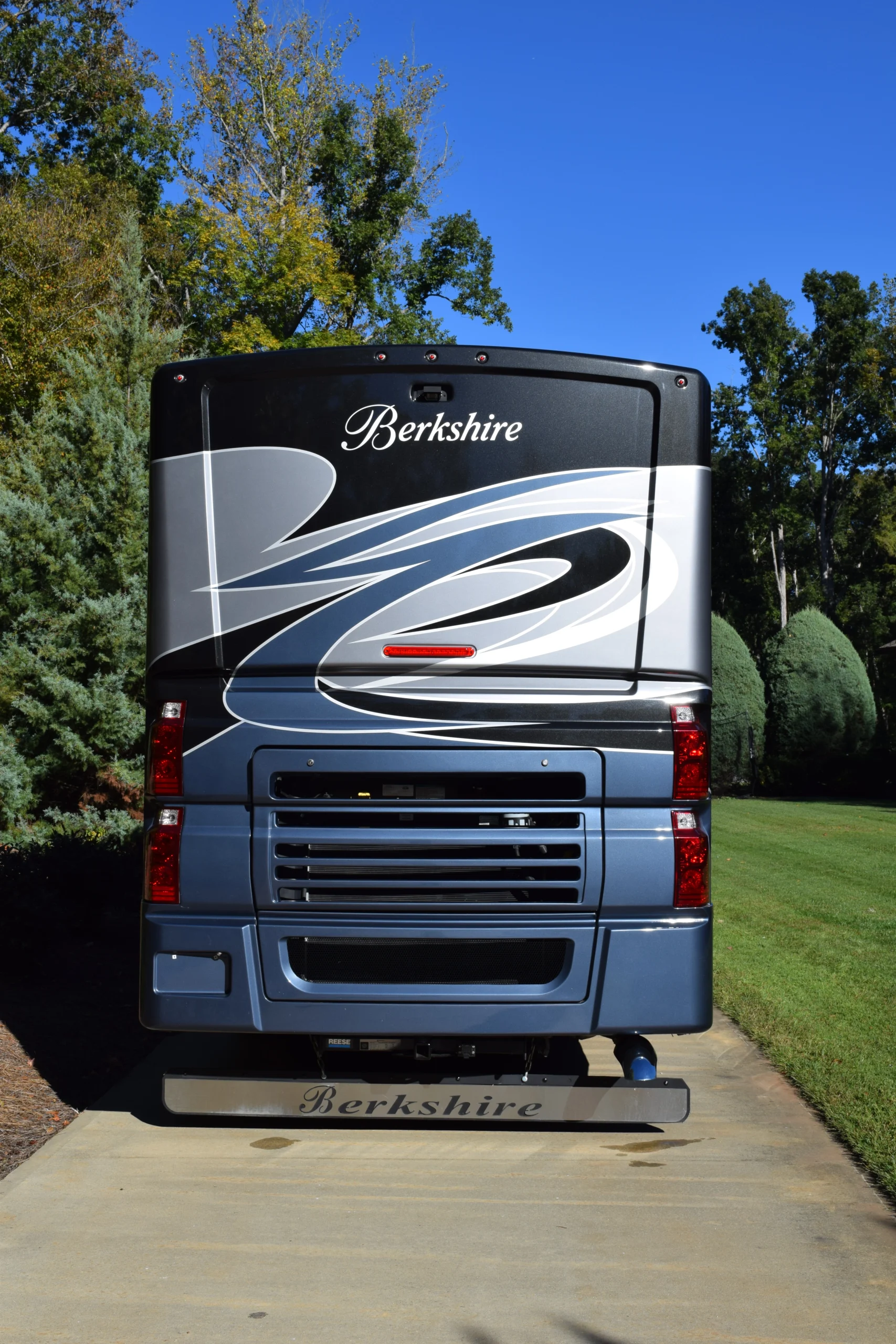 DSC_0693 Rear view of a Berkshire motorcoach with sleek blue and silver design, parked on a driveway, surrounded by lush green grass and trees.