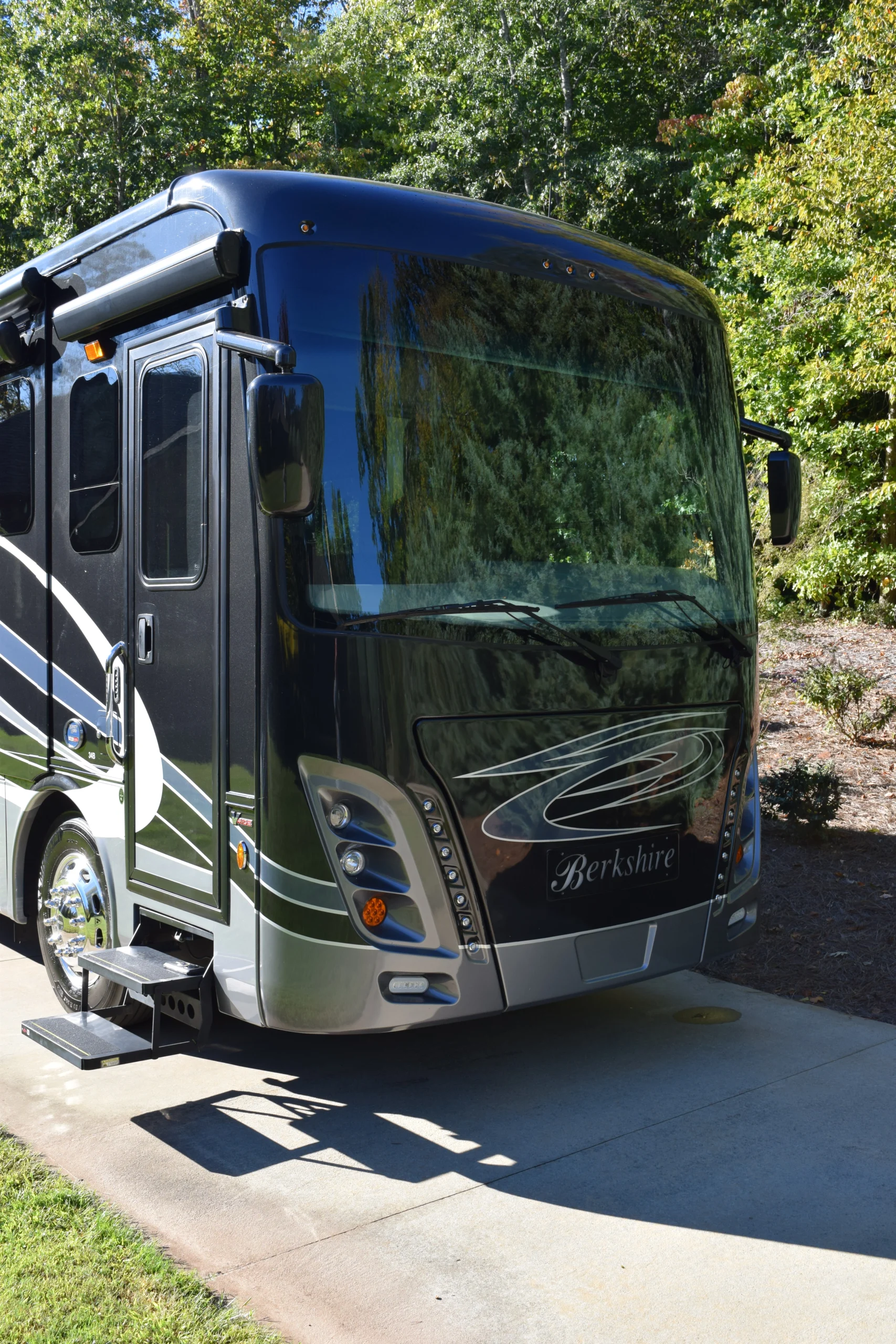 DSC_0694 Luxurious motorcoach parked on a sunny driveway, surrounded by lush green trees, showcasing sleek design and shiny chrome wheels.