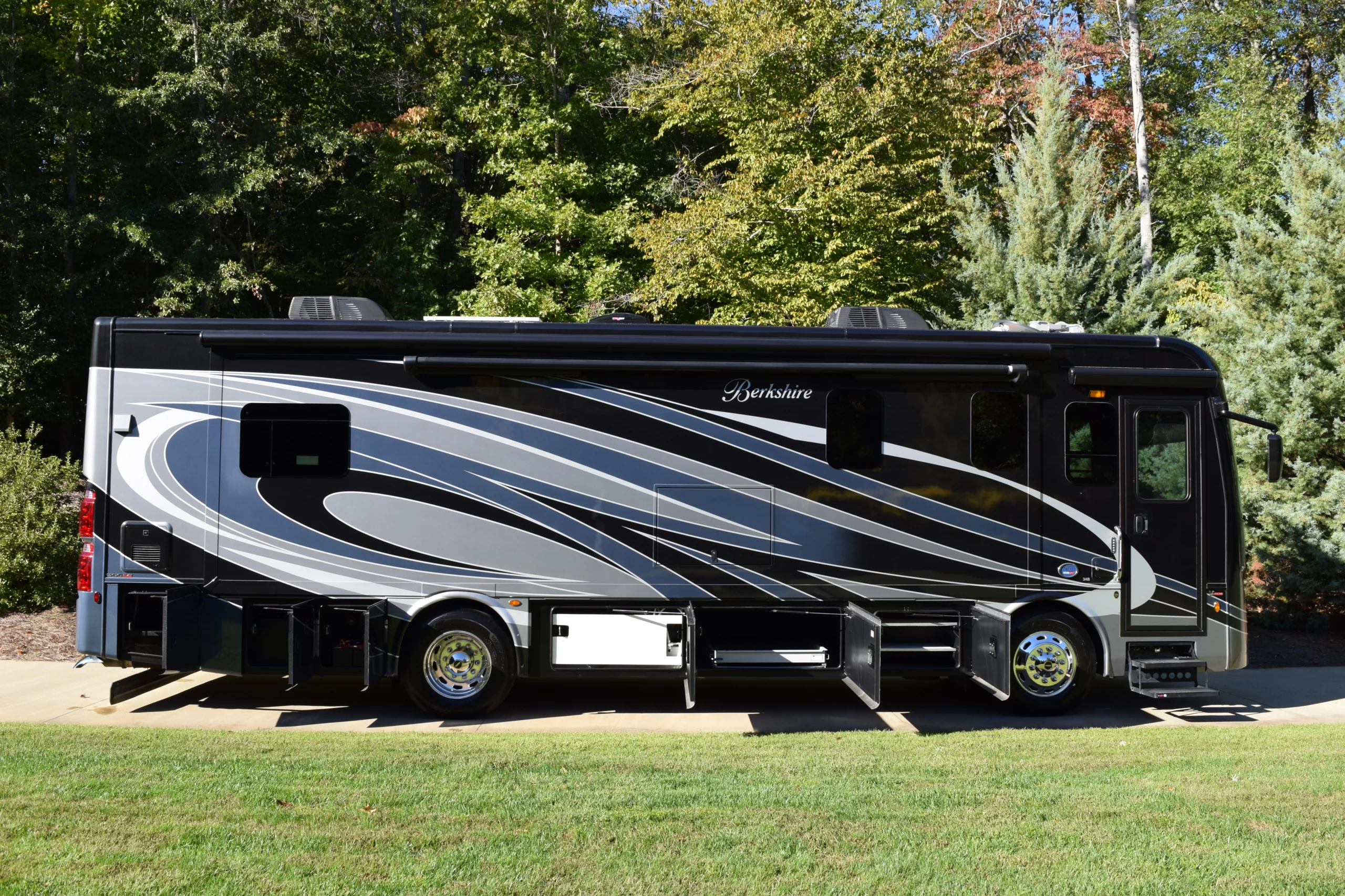DSC_0695 Sleek black and gray motorcoach parked on a concrete driveway with storage compartments open, surrounded by lush greenery.