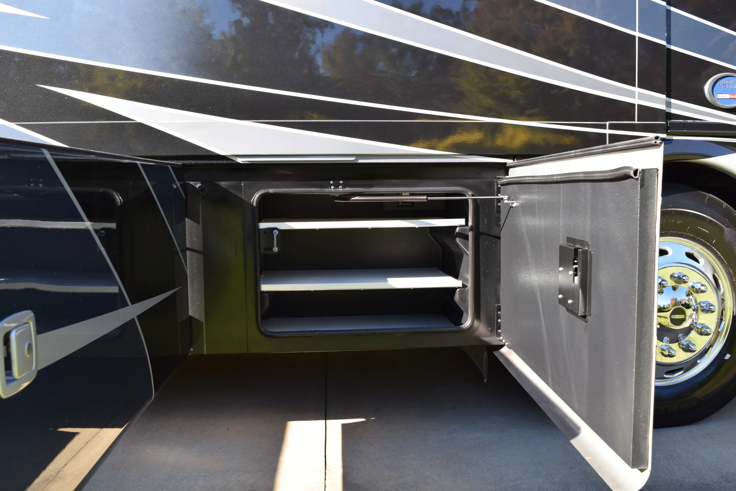 DSC_0697 Open storage compartment with shelves on a Motorcoach and RV, glossy black and silver exterior detailing, reflecting sunlight.