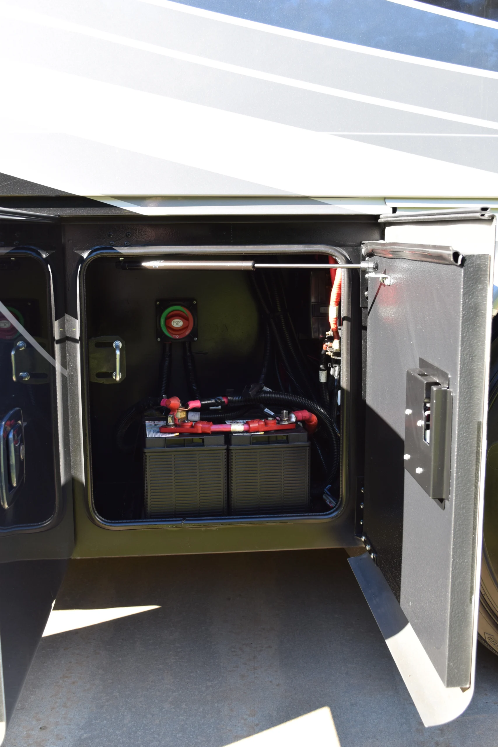 DSC_0701 Open compartment of a motorcoach revealing dual batteries and wiring, door ajar for inspection or maintenance.