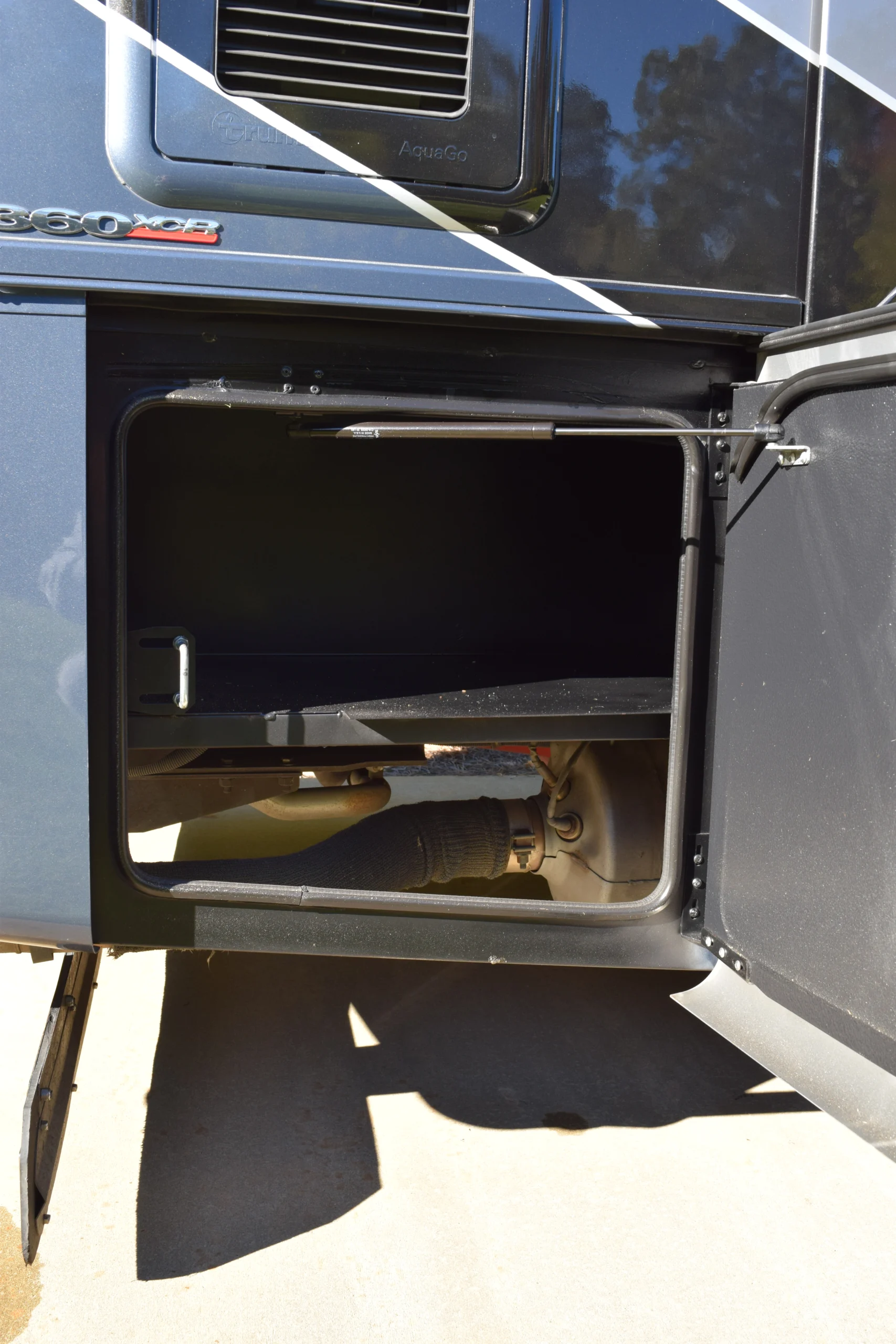 DSC_0703 Open storage compartment on a Motorcoach and Rvs, revealing vehicle’s exhaust system and a shelf inside.