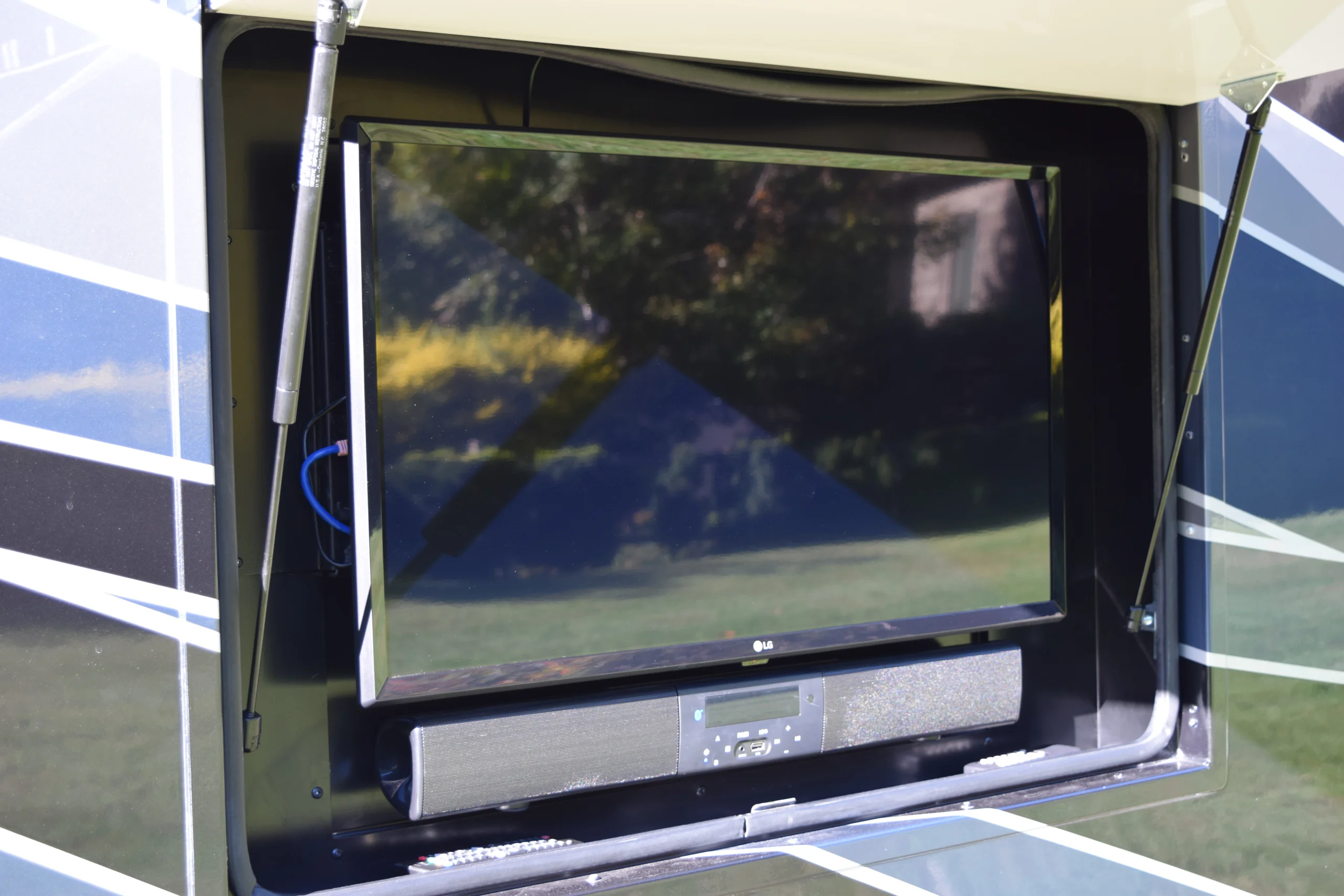 DSC_0710 Outdoor TV with soundbar installed in a motorcoach's storage compartment, reflecting greenery.