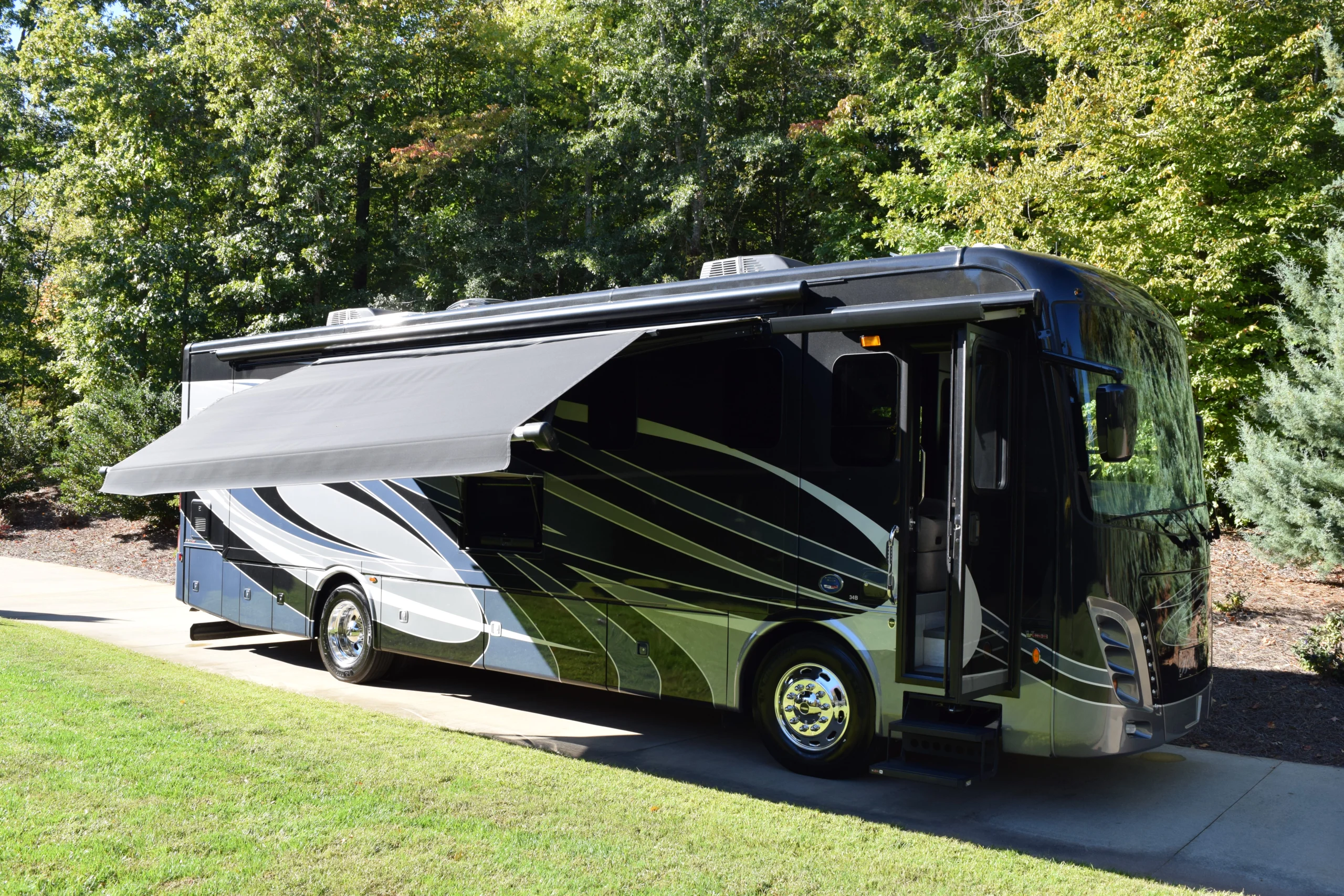 DSC_0711 Sleek motorcoach parked on a driveway, extended awning, surrounded by lush green trees.