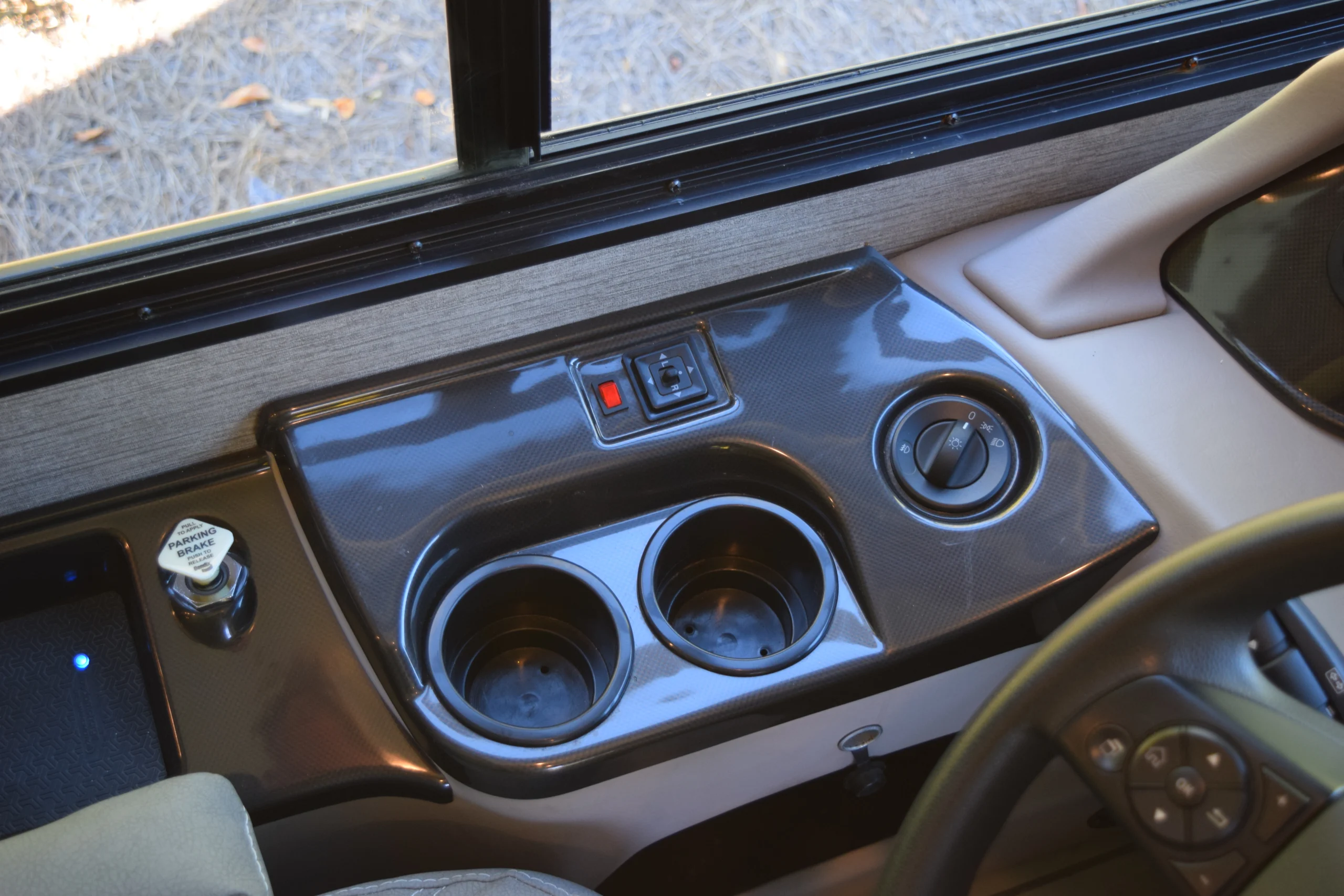 DSC_0717 Dashboard of a motorcoach with dual cup holders, parking brake, and control knobs near window.