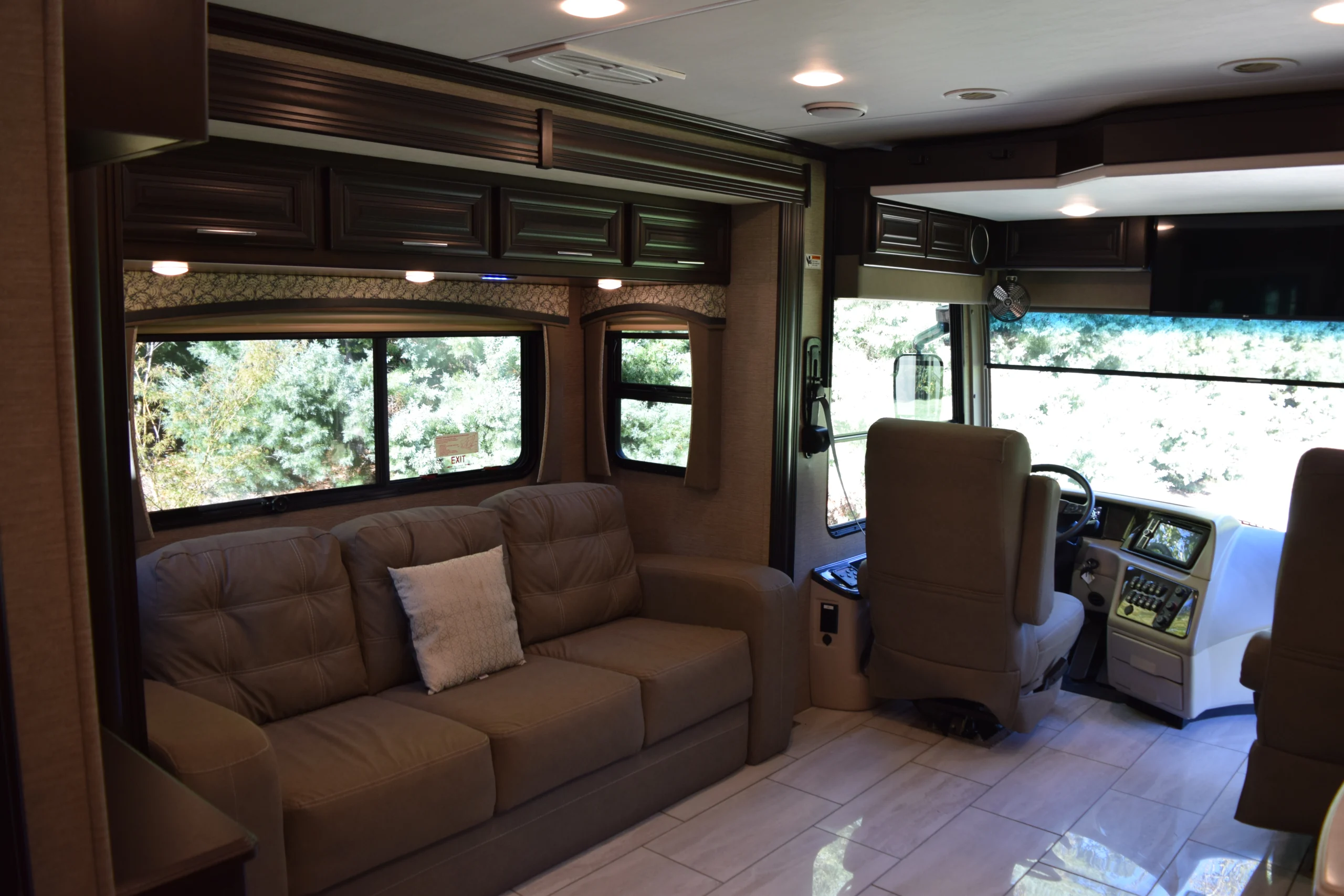 DSC_0718 Interior of a motorcoach showing a plush brown sofa, driver’s cabin, large windows, and overhead cabinets, with a serene outdoor view.