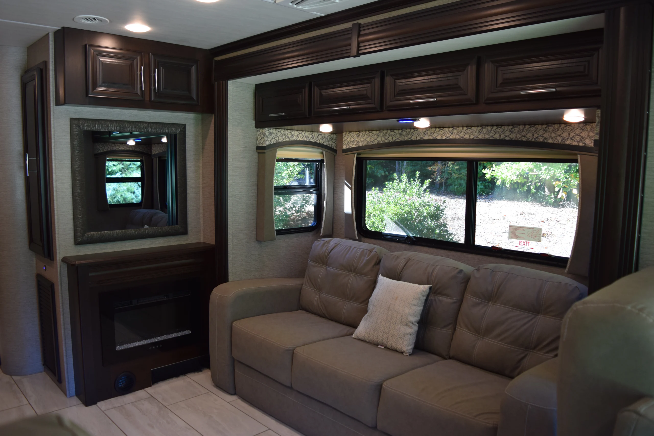DSC_0719 Spacious Motorcoach RV living area with a large sofa, dark wood cabinets, fireplace, and window view of greenery.