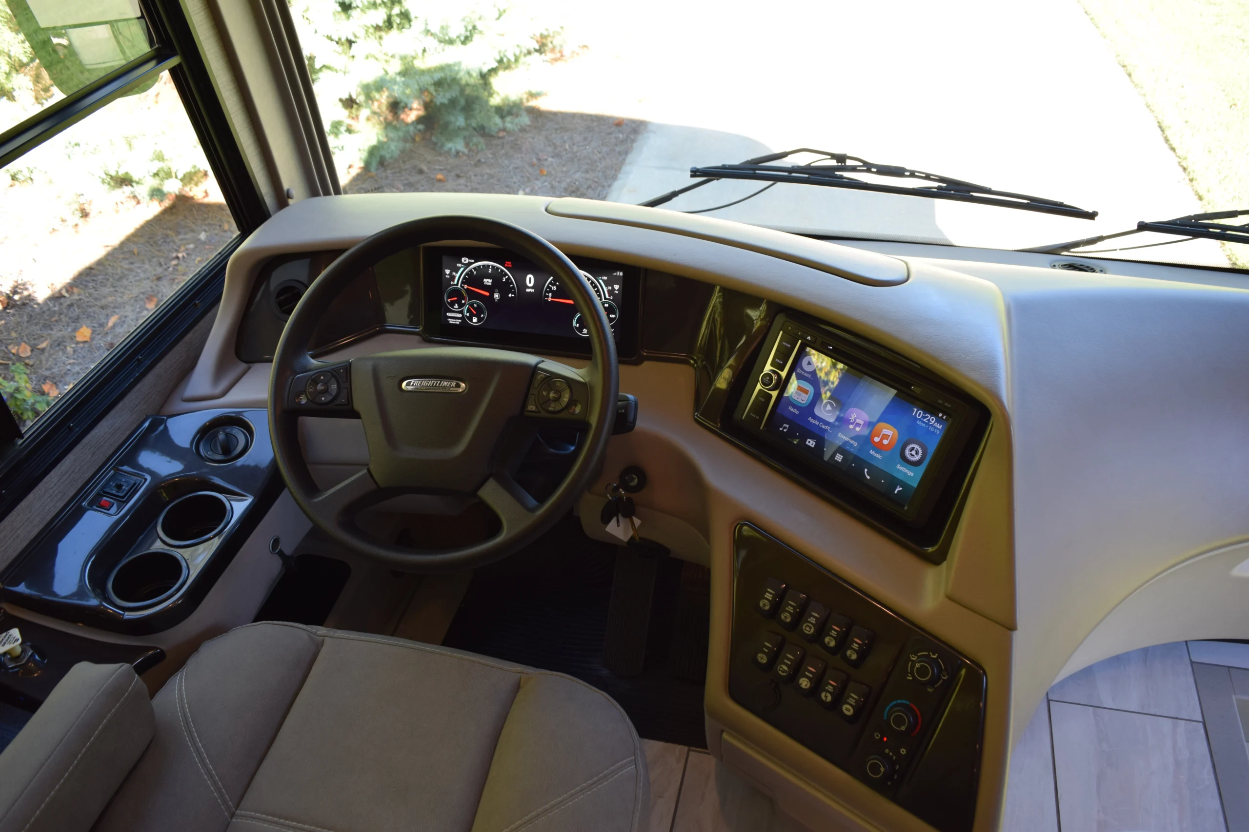 DSC_0720 Motorcoach driver's cockpit with steering wheel, digital dashboard, control panel, and touchscreen display, overlooking a driveway.