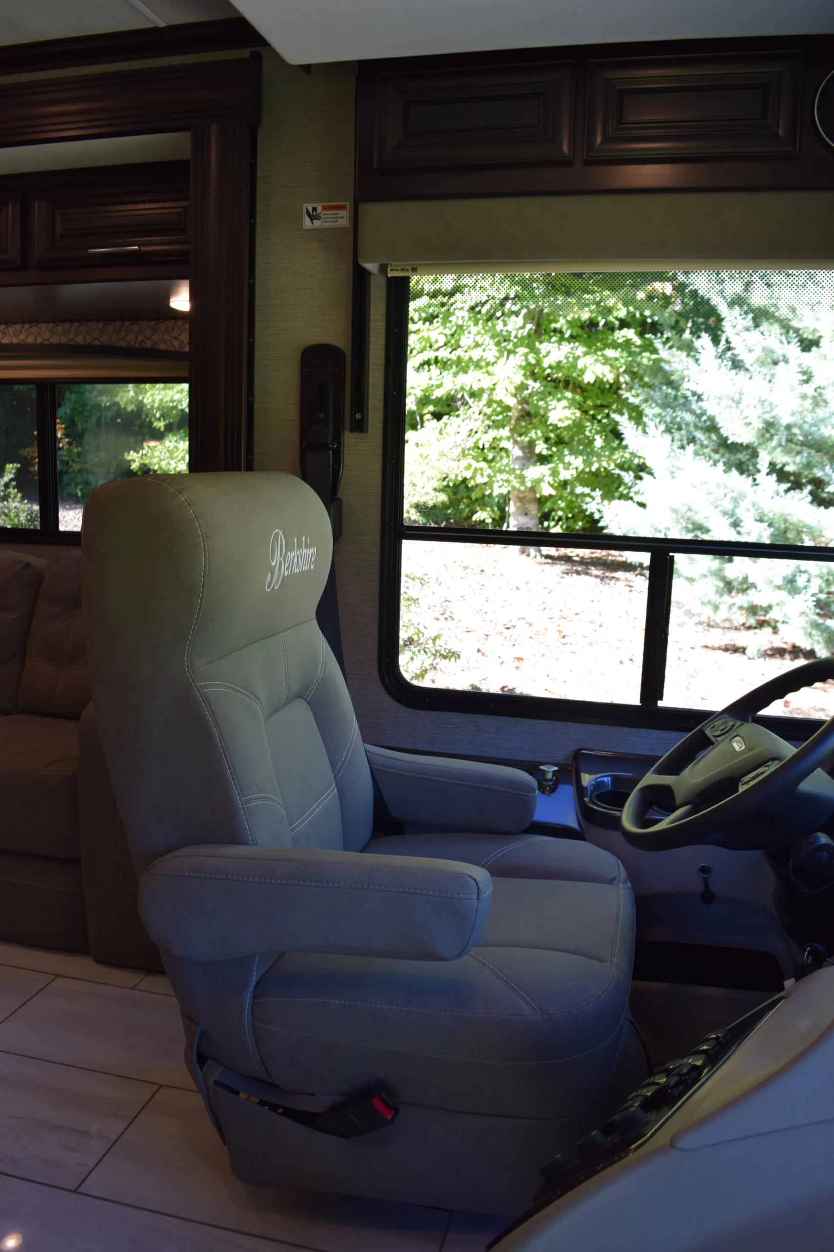 DSC_0721 Motorcoach and RVs driver seat with beige upholstery, adjacent steering wheel, and large window showing greenery outside.