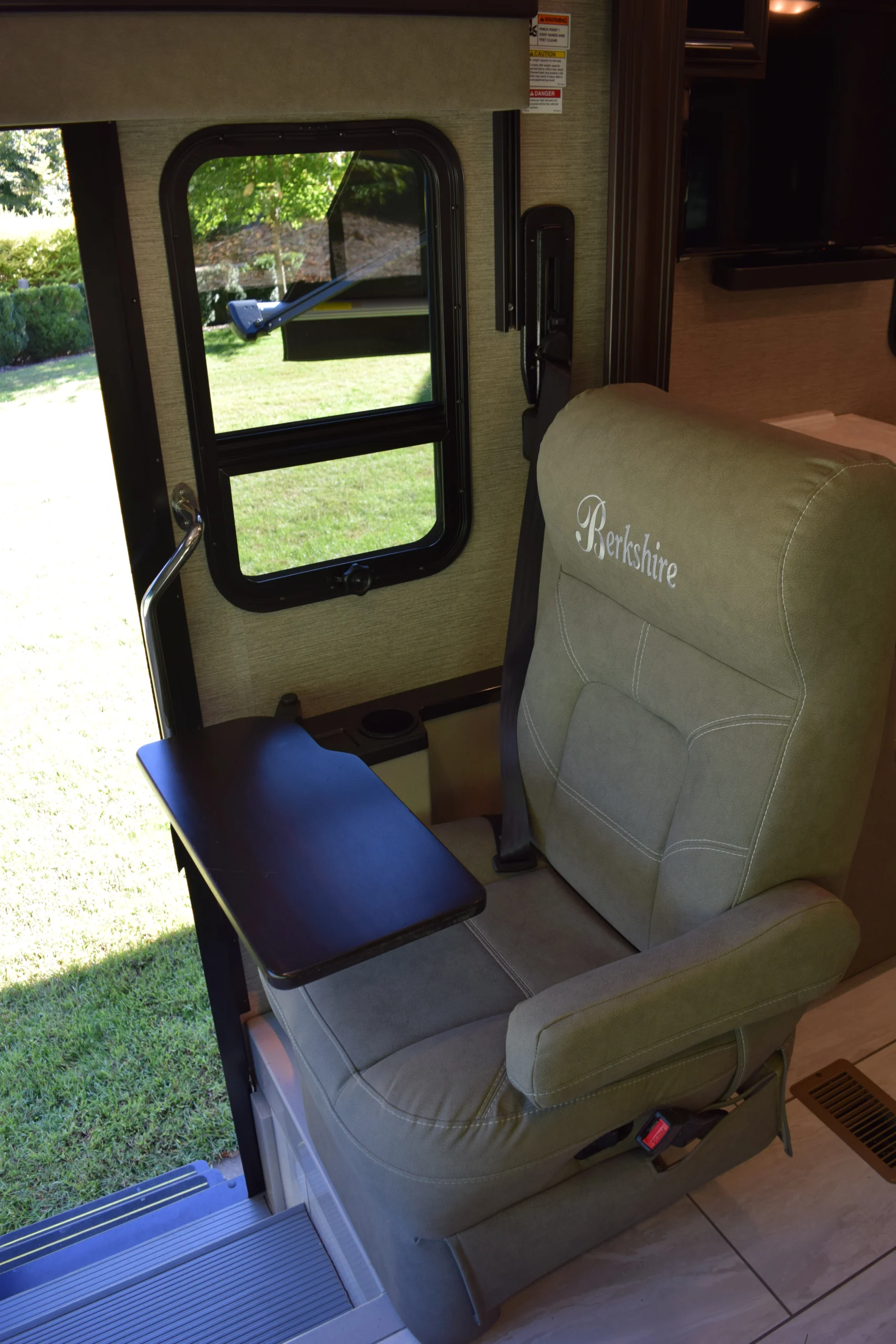 DSC_0723 Motorcoach interior with a beige captain's chair labeled "Berkshire," a foldable tray, and an open door showing grass outside.