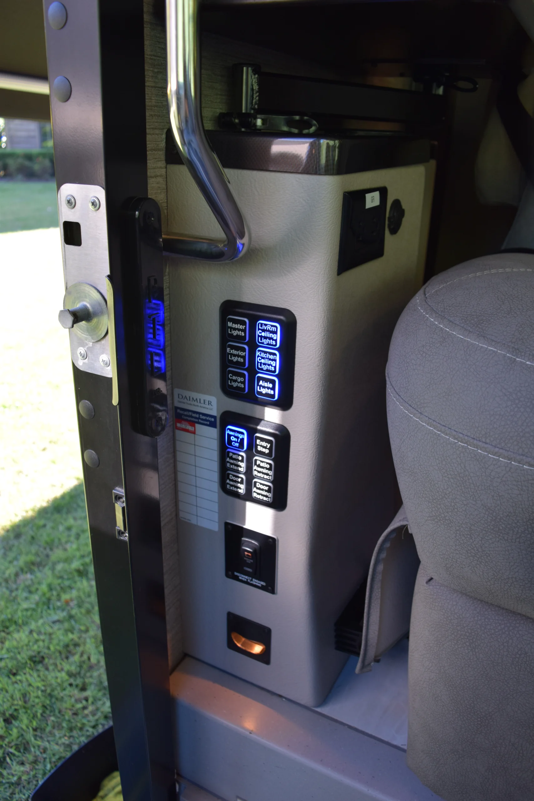 DSC_0725 Control panel with illuminated buttons for lights and entry steps in a motorcoach, near a leather seat and metal handrail.