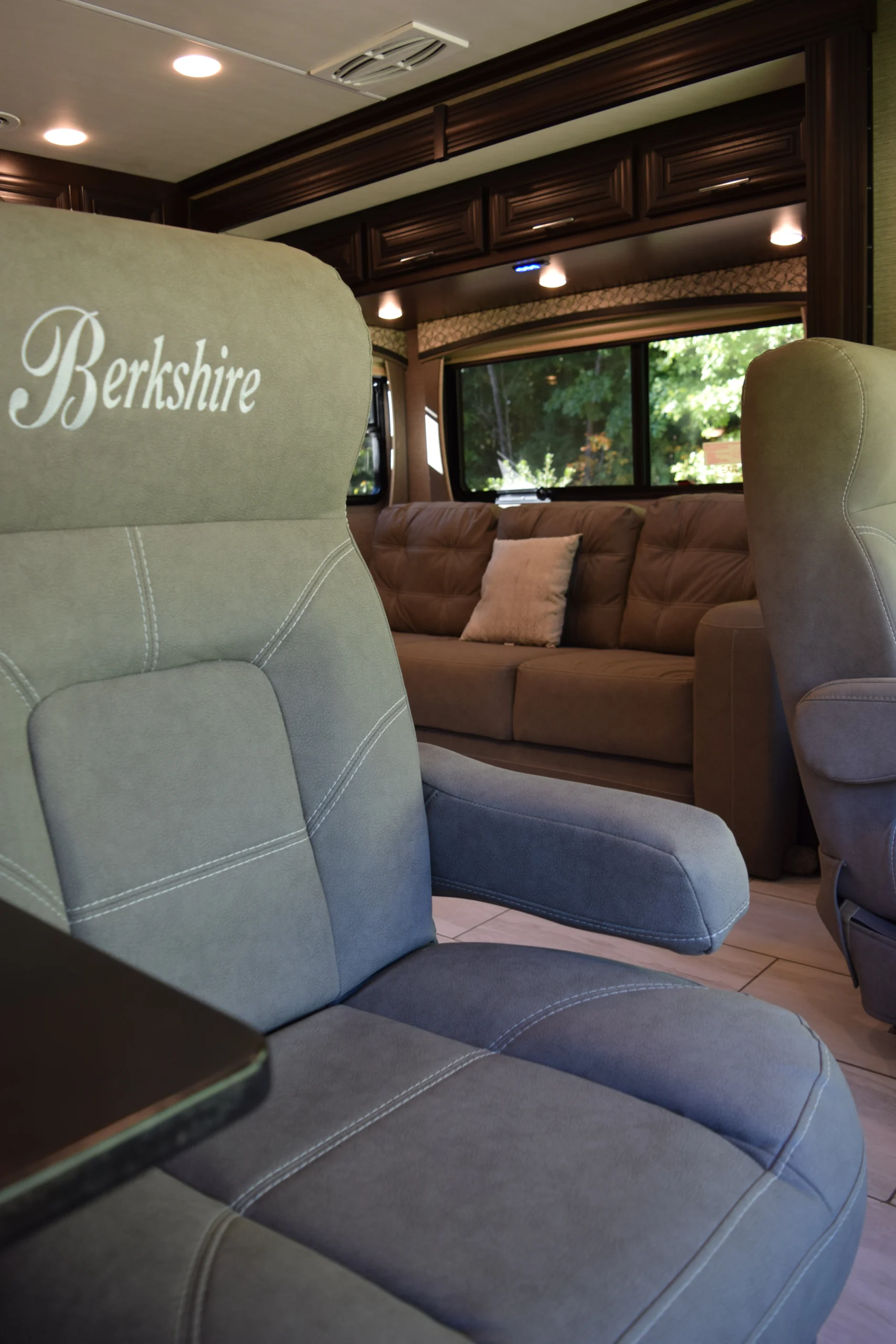 DSC_0726 Luxury motorcoach interior with a "Berkshire" embroidered captain's chair, plush brown sofa, wooden cabinets, and large windows.