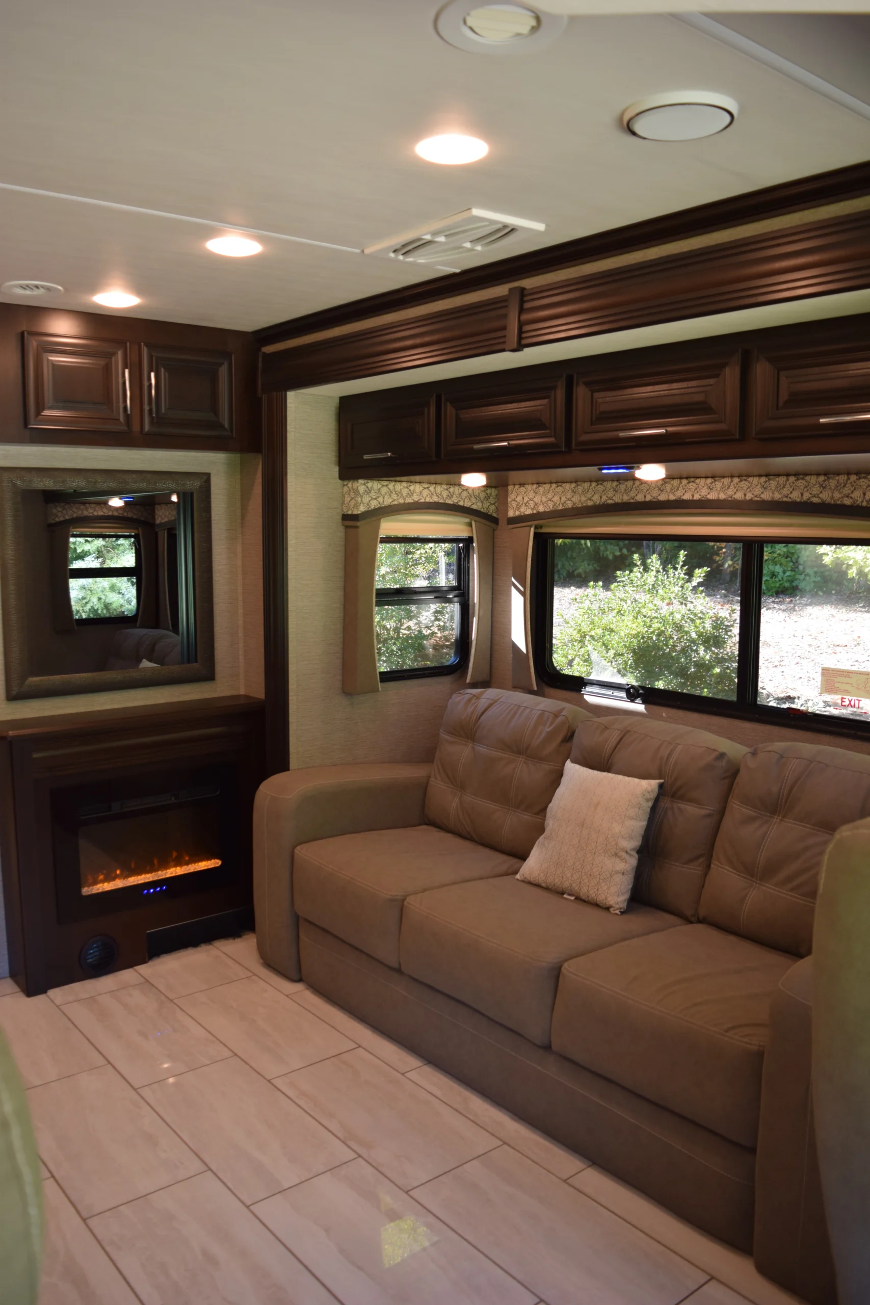 DSC_0727 Cozy motorcoach interior with a brown sofa, decorative pillow, electric fireplace, mirror, and large window showing greenery outside.