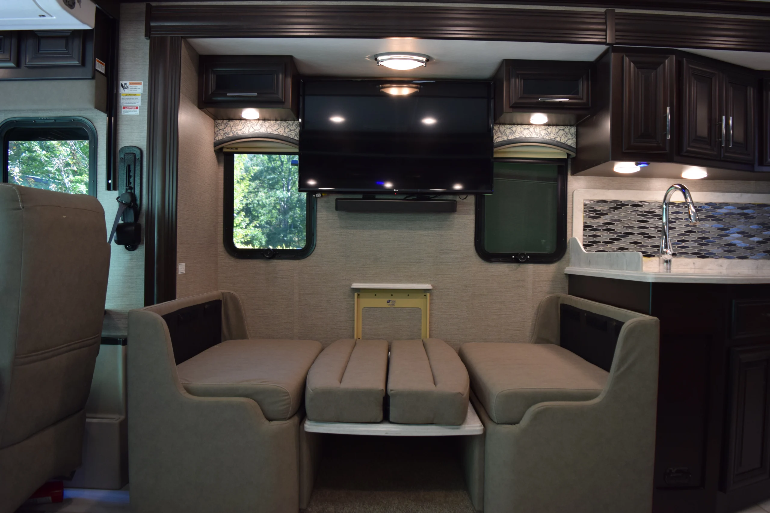 DSC_0732 Cozy Motorcoach and Rvs interior with gray couches, wall-mounted TV, and kitchenette with modern backsplash and dark wood cabinets.