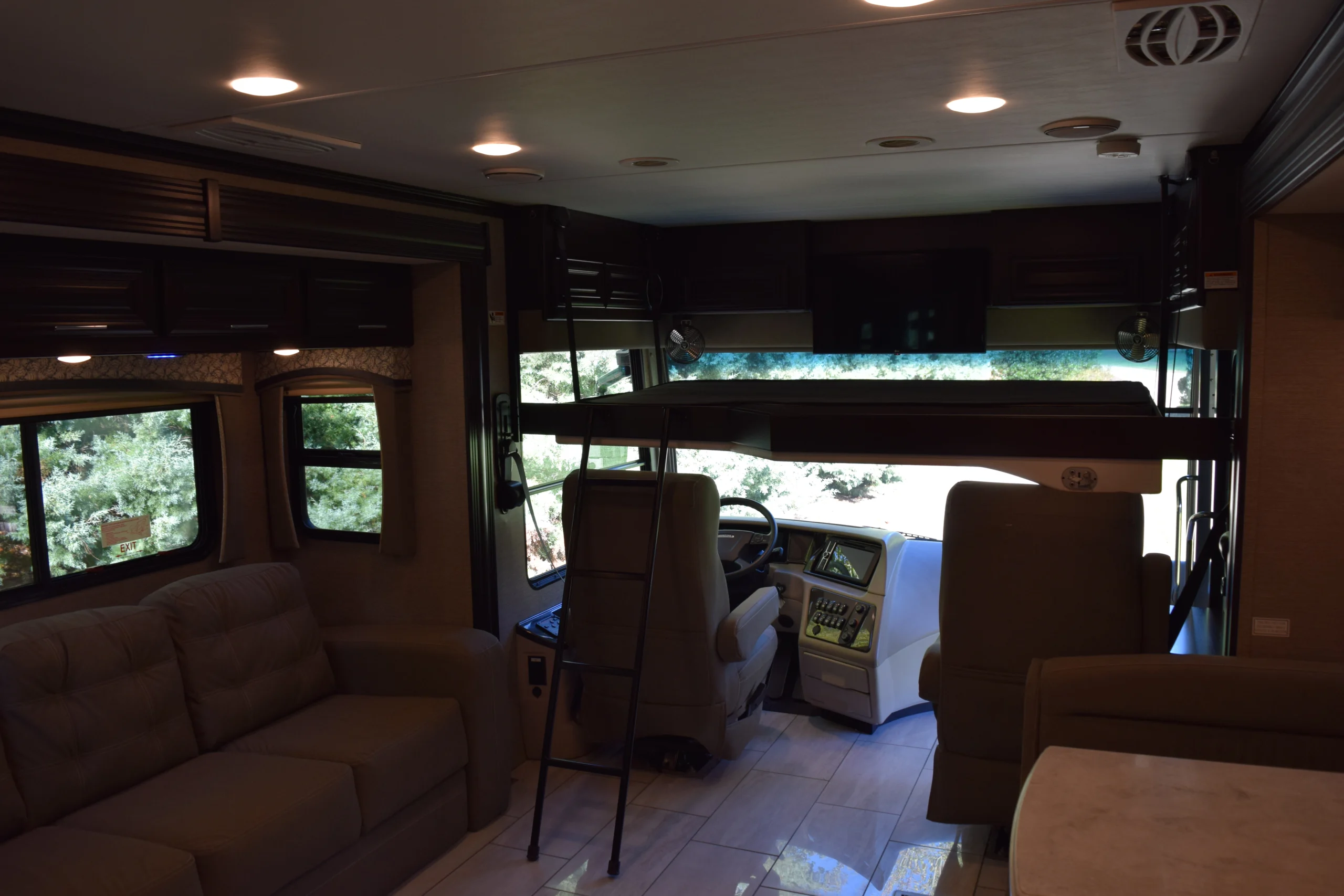 DSC_0734 Interior of a motorcoach with beige seating, a steering area, large windows, overhead storage, and a small ladder leading to a bunk.