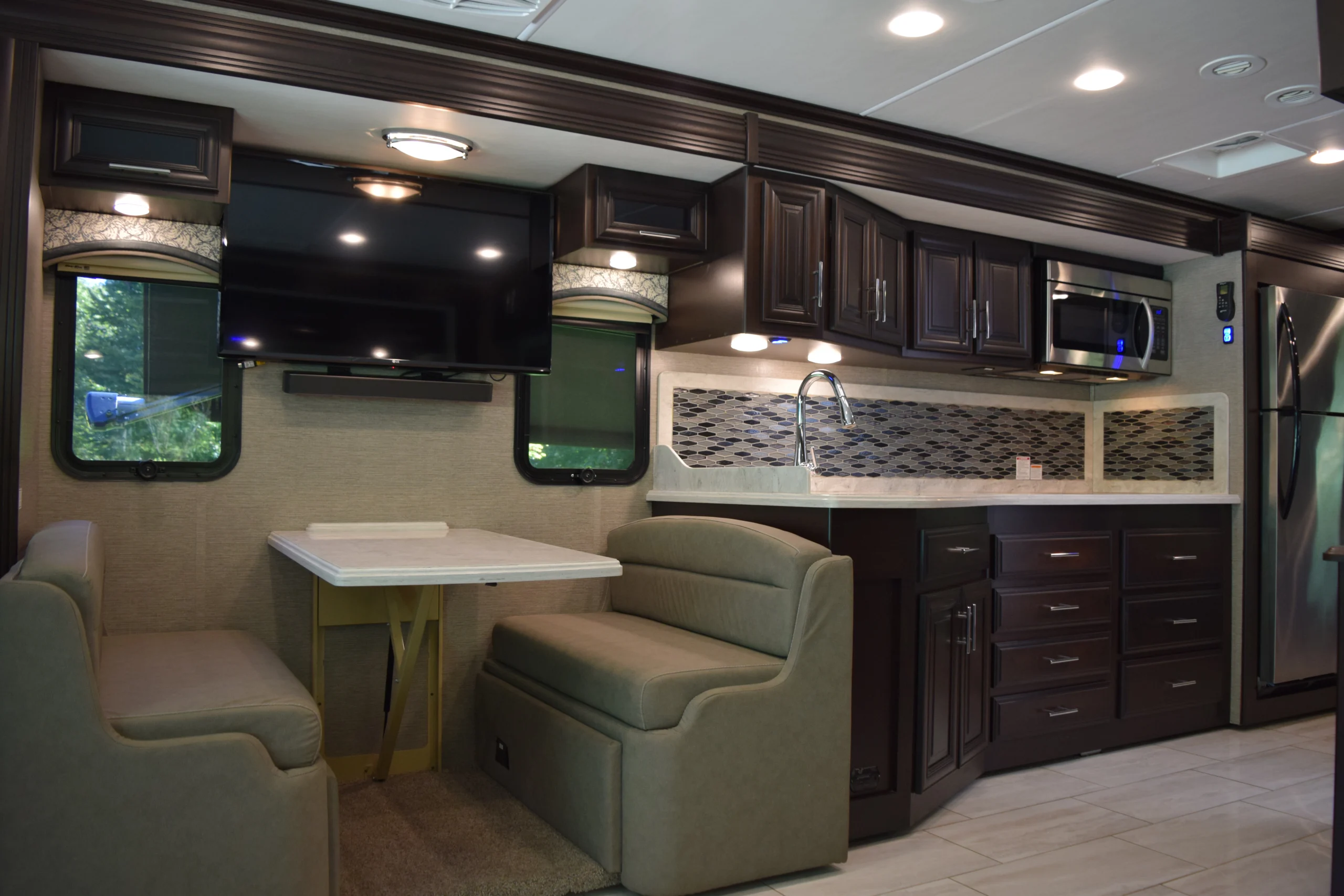 DSC_0736 Luxurious Motorcoach interior with dark wood cabinets, a dining area, TV, and modern kitchen featuring mosaic backsplash and stainless appliances.