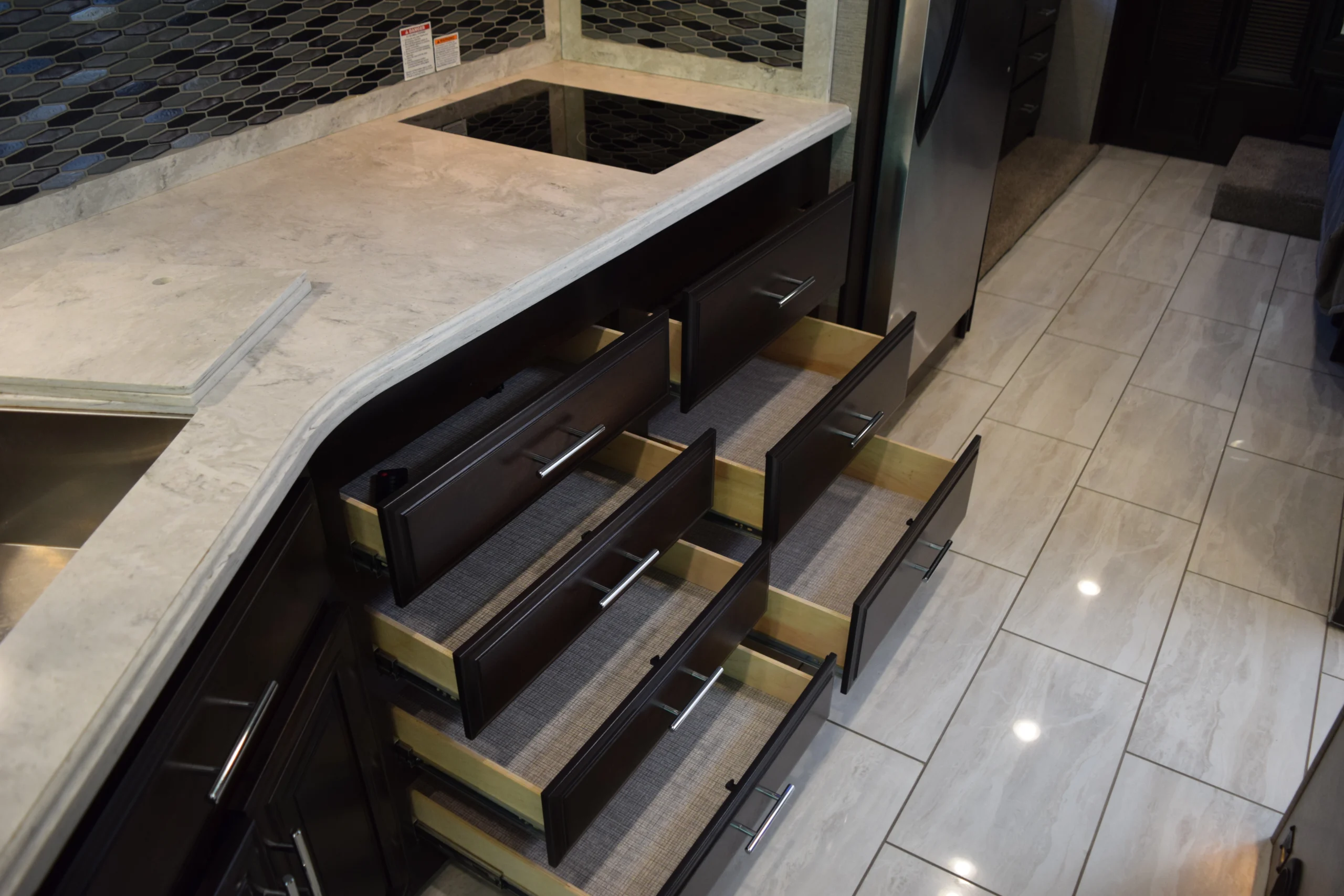 DSC_0741 RV kitchen with marble countertop, open dark wood drawers, stainless steel fridge, and tile floor.