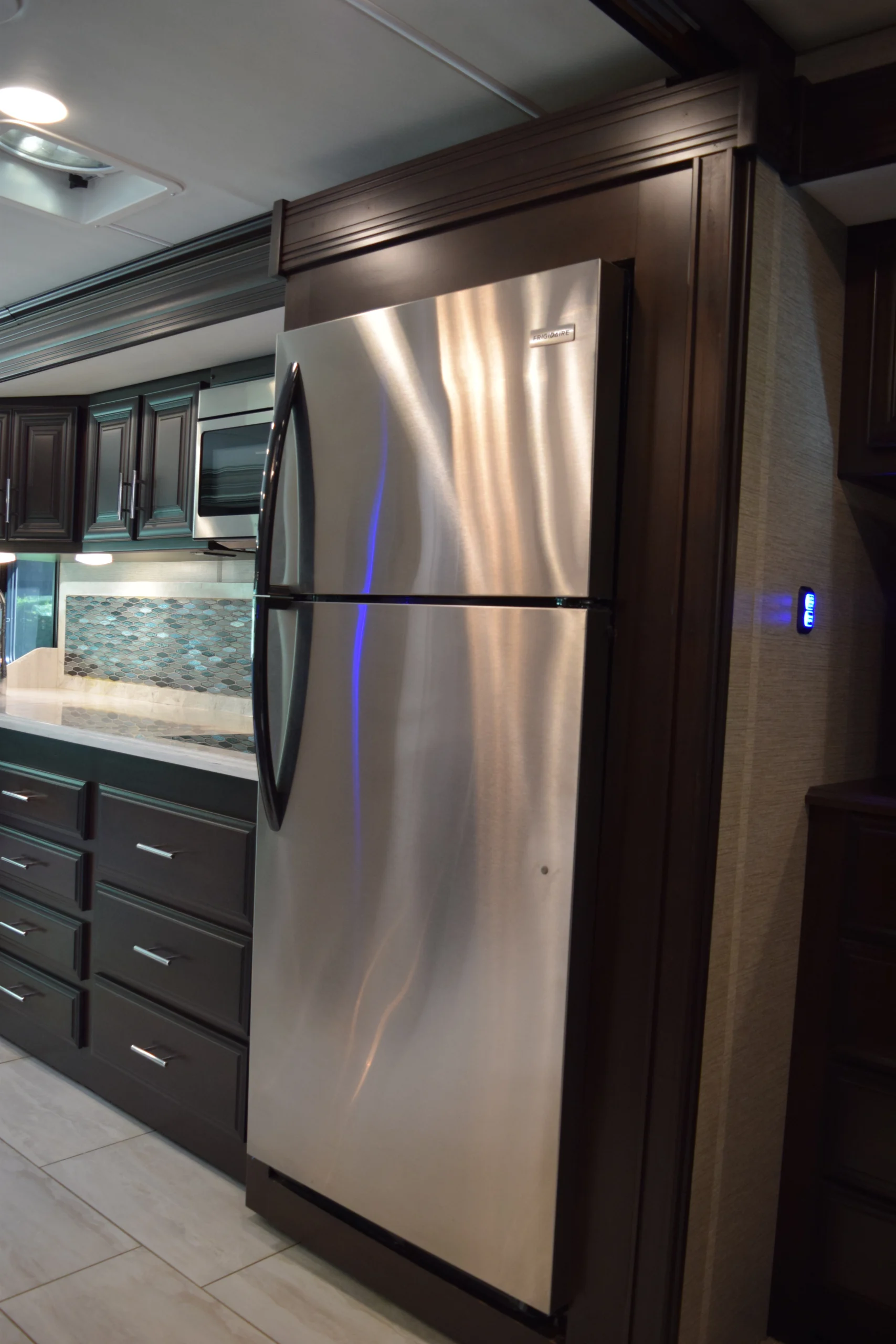 DSC_0744 Motorcoach kitchen with stainless steel fridge, dark wooden cabinets, modern backsplash, and under-cabinet lighting.
