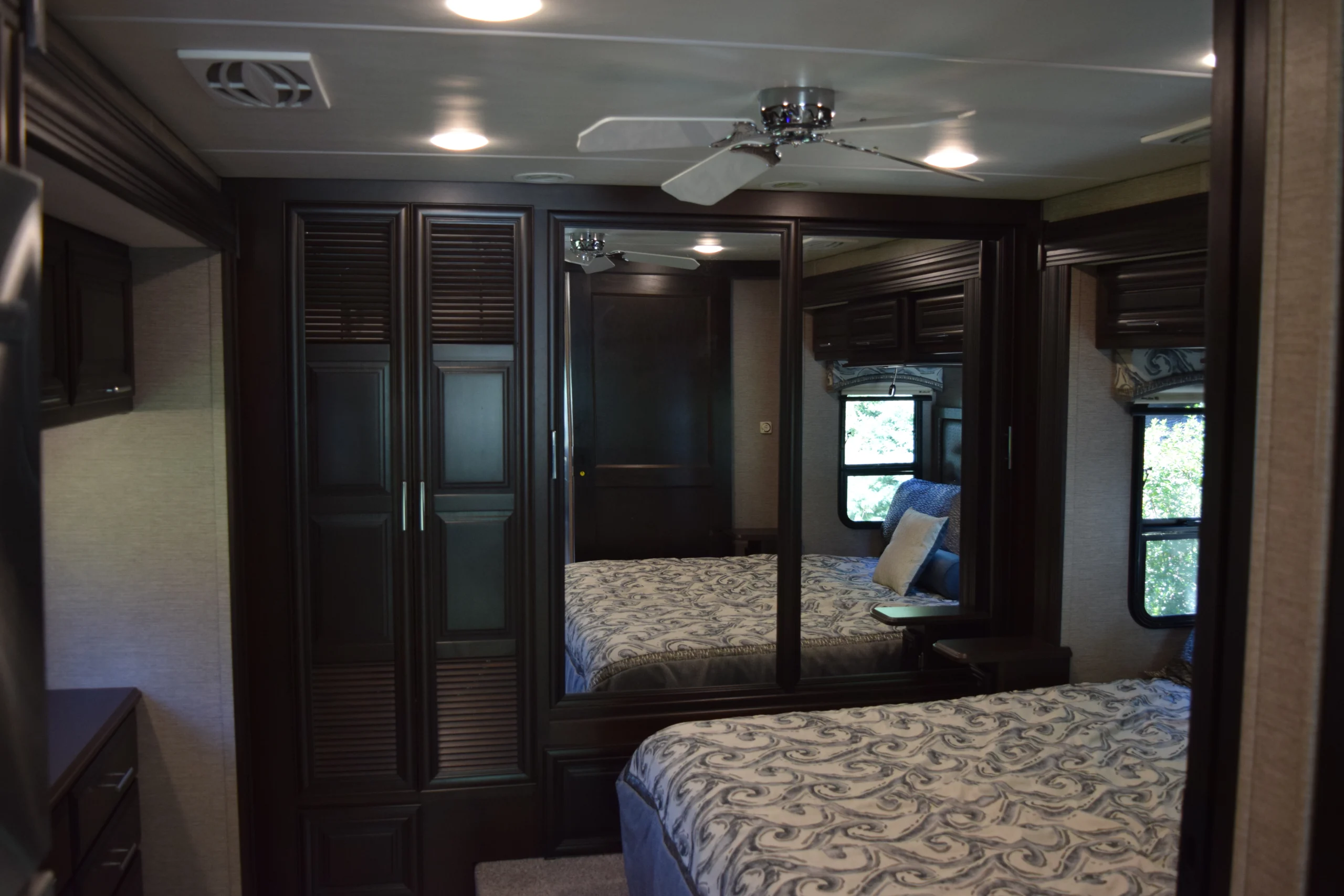 DSC_0747 Luxurious motorcoach and RV bedroom with dark wood cabinets, mirrored closet doors, ceiling fan, and patterned bedding.