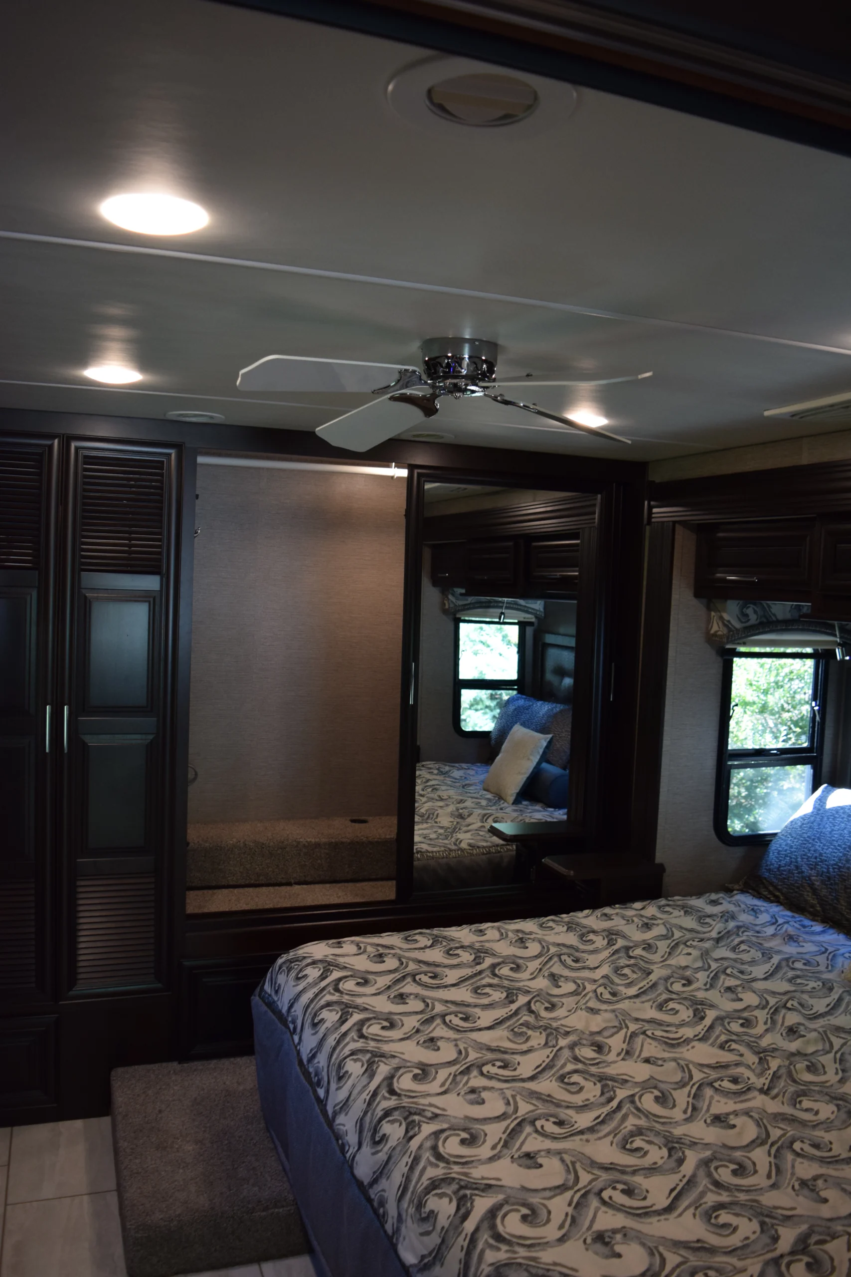 DSC_0749 Luxurious Motorcoach bedroom with ceiling fan, ornate bedding, dark wood cabinets, and mirrored closet reflecting windows with outdoor view.