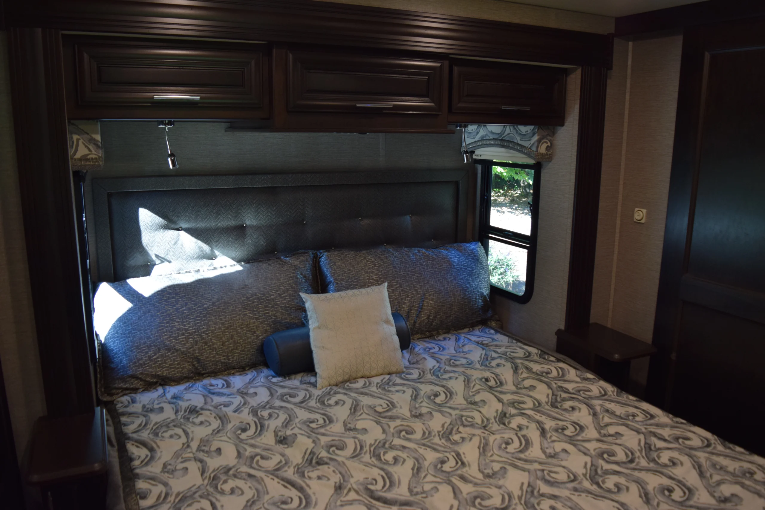 DSC_0750 Luxurious RV bedroom with plush pillows, decorative bedding, dark wood cabinetry, and sunlight through the window. Perfect for Motorcoach and RVs.