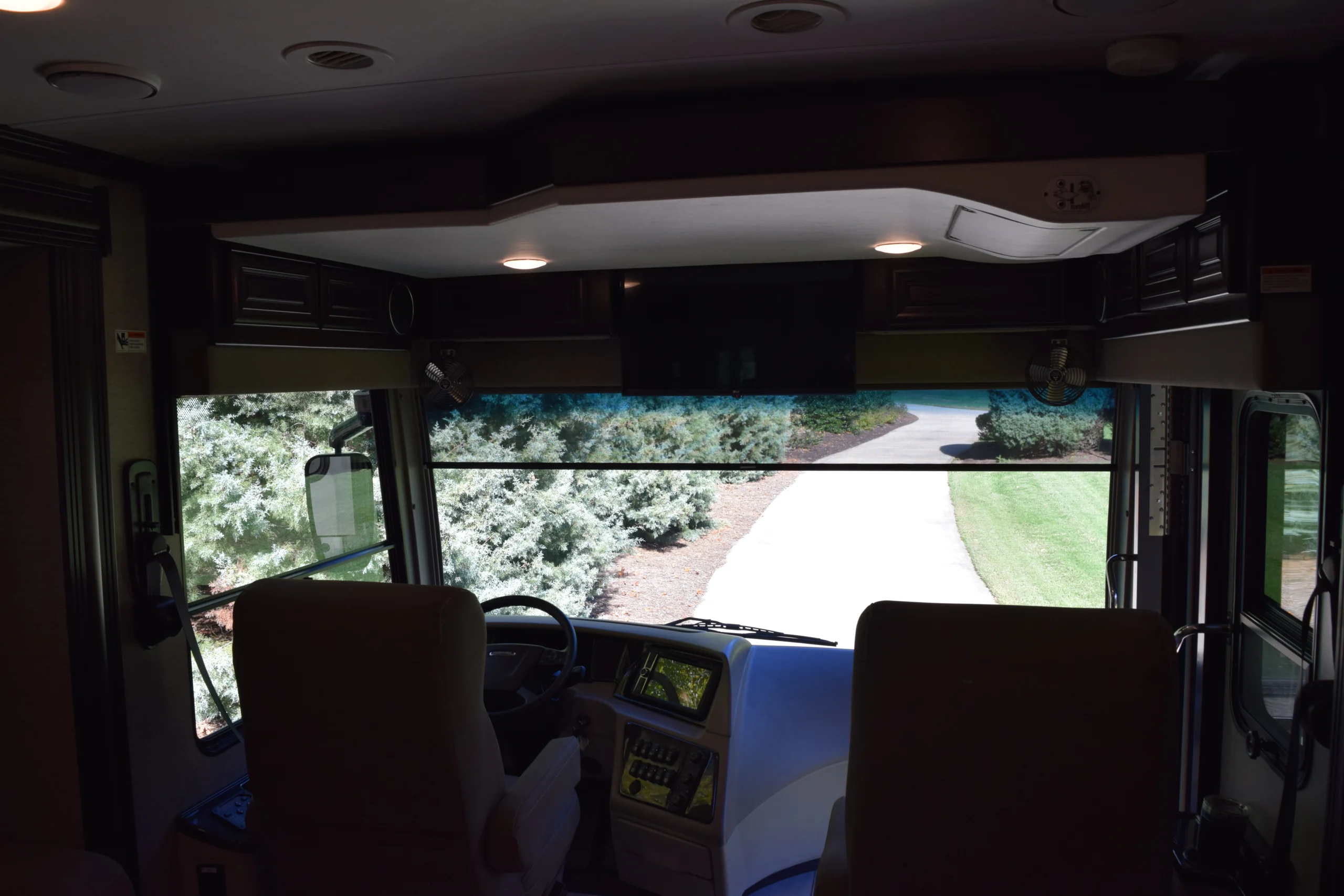 DSC_0755 Motorcoach interior with driver seats, steering wheel, and dashboard, facing a clear windshield view of a garden-lined driveway.