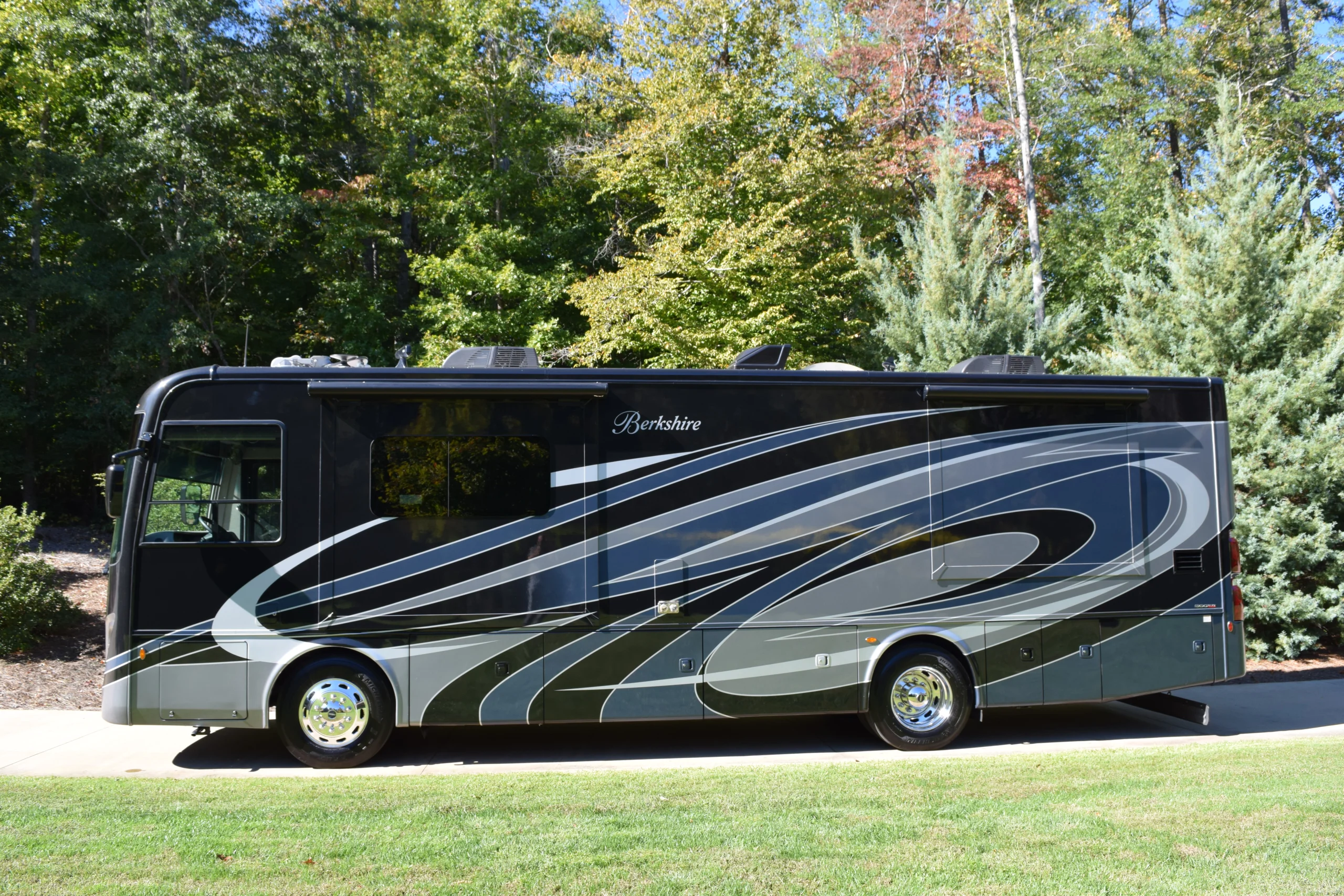 DSC_0766 Sleek black motorcoach with silver swirls parked on a sunny day, surrounded by lush green trees.