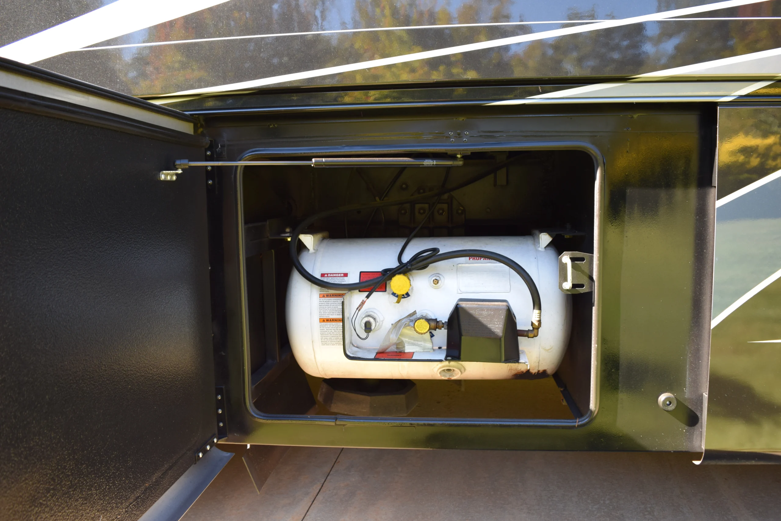 DSC_0767 Propane tank compartment on a Motorcoach RV with open hatch, revealing a secured white tank and connecting hose.
