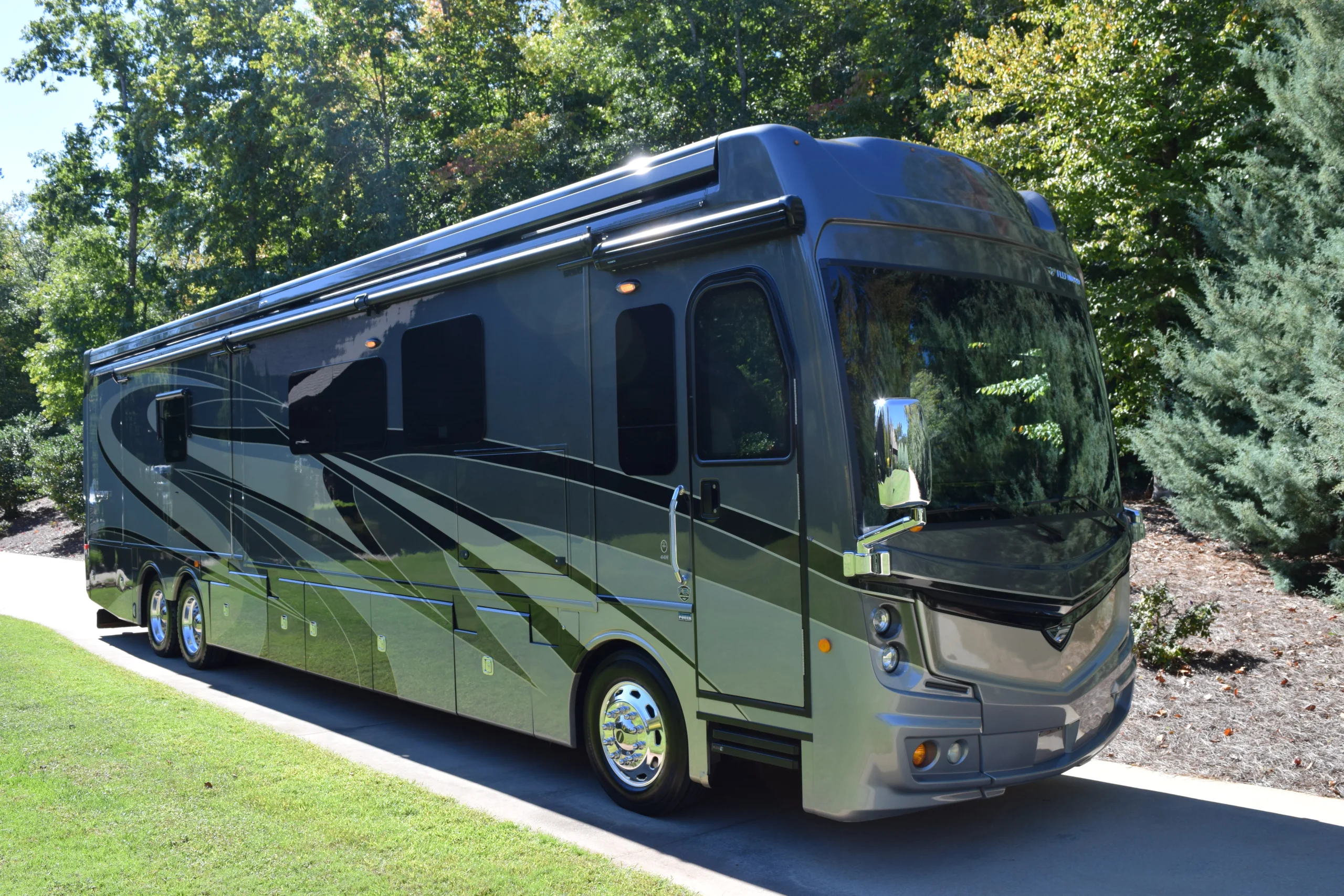 Sleek, dark grey motorcoach parked on a driveway surrounded by lush green trees, showcasing its reflective windows and modern design.