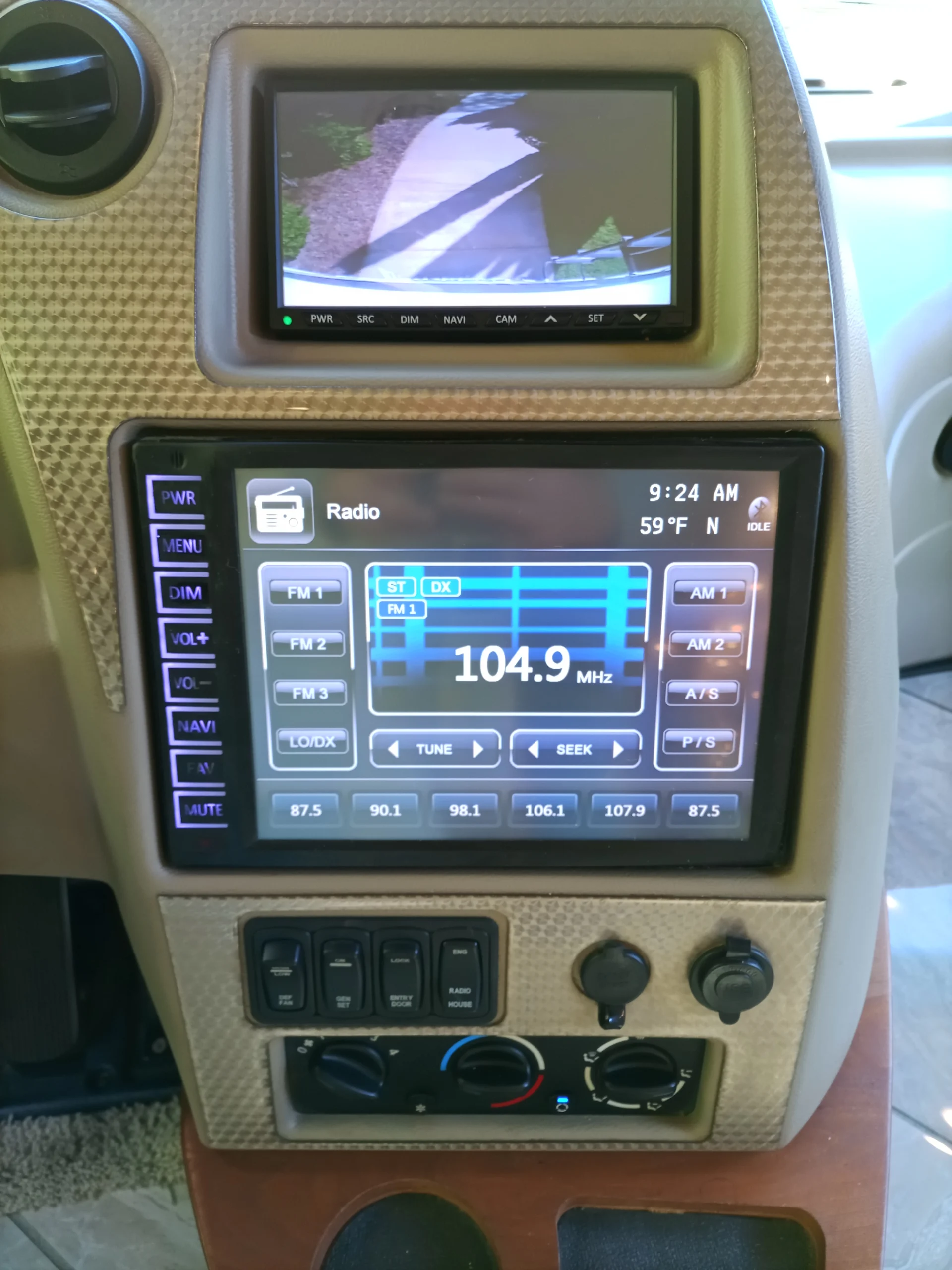 IMG_20260928_092406_206 Motorcoach and RVs dashboard with a backup camera display, radio screen showing 104.9 MHz, climate controls, and various buttons.