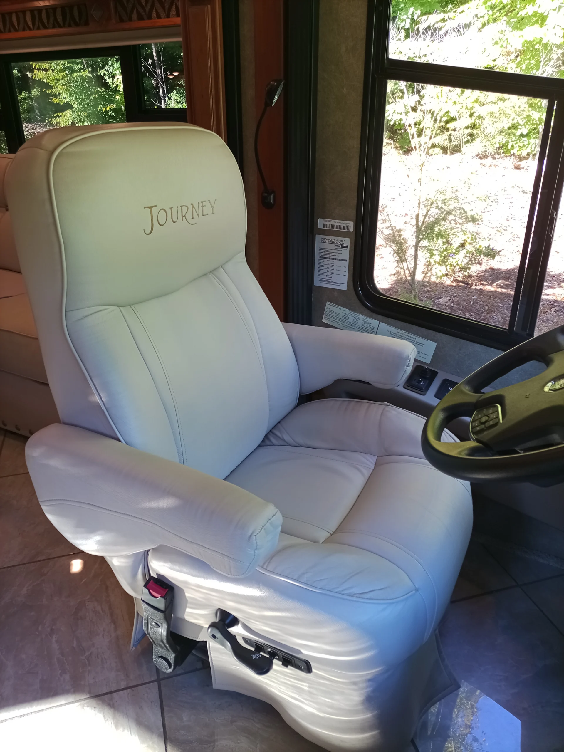 IMG_20260928_092438_432 White leather driver's seat in a motorcoach with "Journey" on the headrest, near window showing greenery outside.