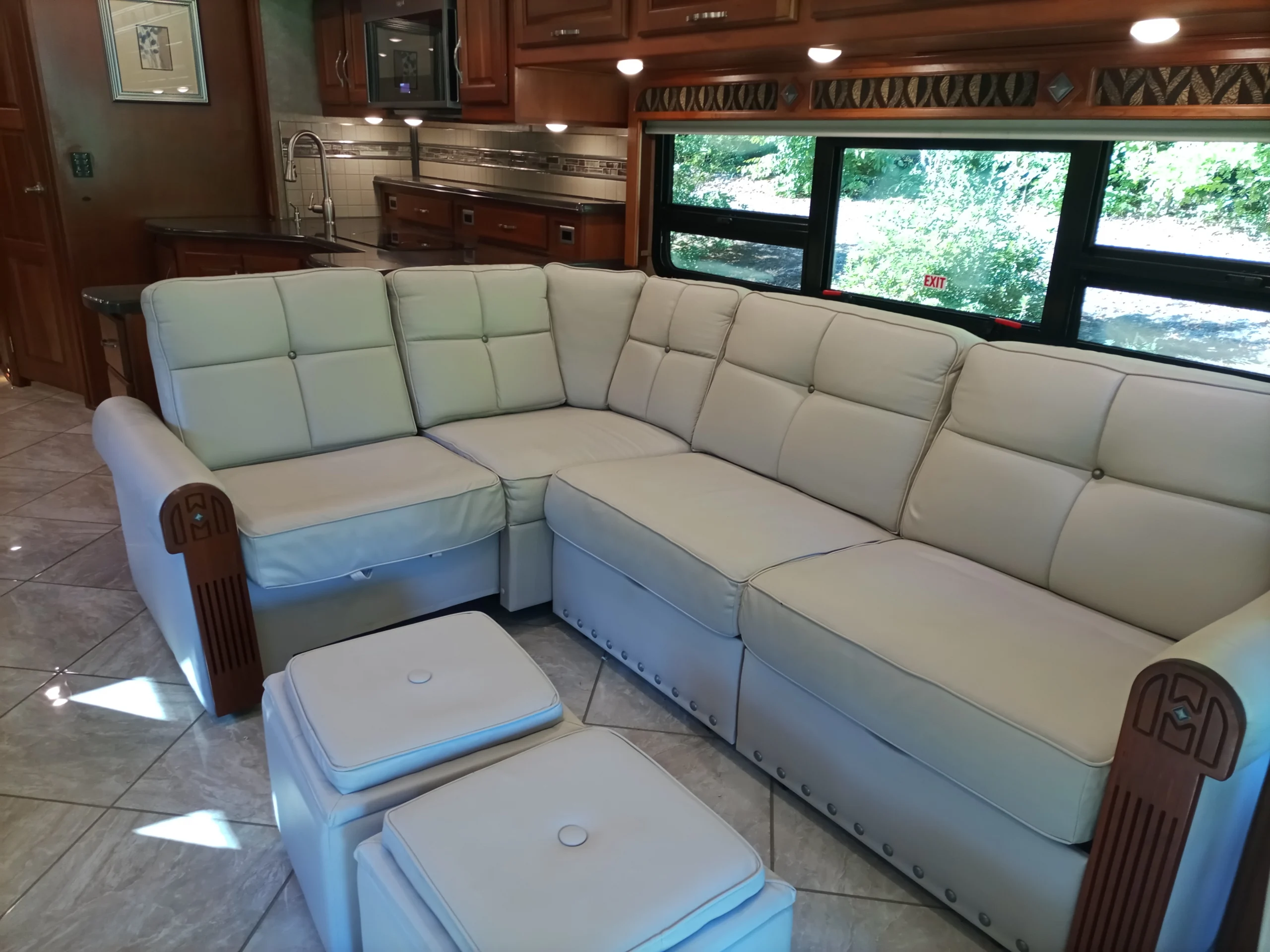 IMG_20260928_092445_586 Luxurious L-shaped beige sofa in a motorcoach, with ottomans, wooden accents, and a view of greenery through large windows.