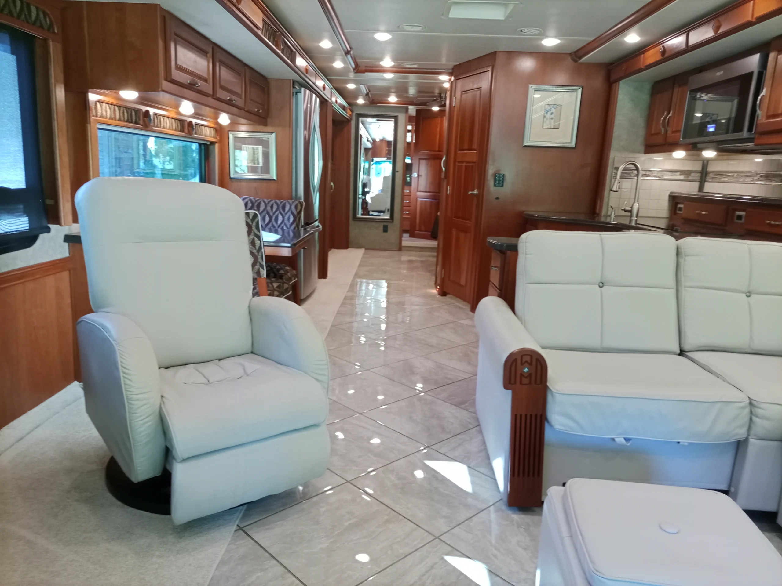 IMG_20260928_092623_509 Luxurious motorcoach interior with white lounge chairs, wooden cabinets, tiled floors, and modern kitchen.