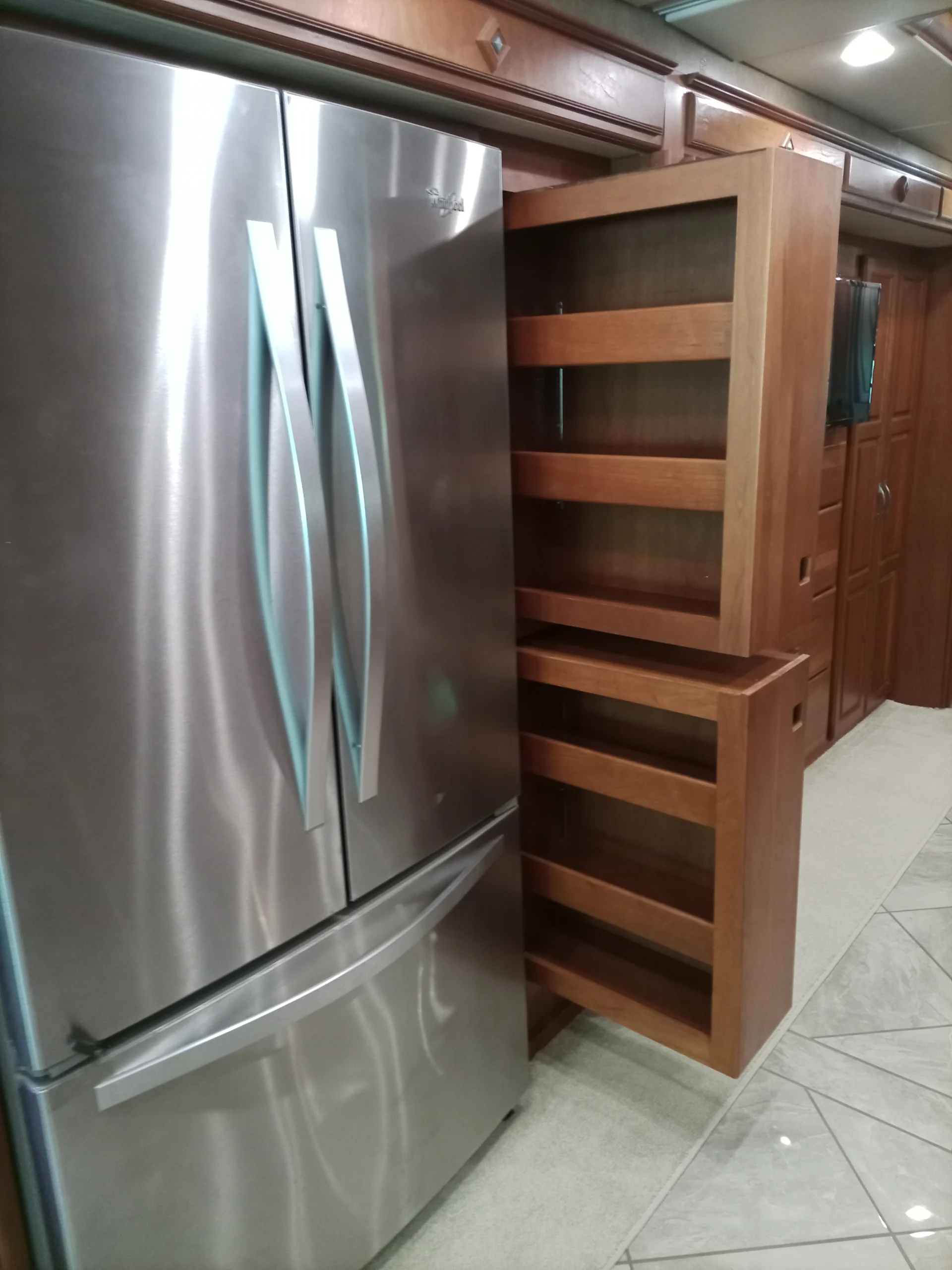 IMG_20260928_092837_615 Stainless steel refrigerator next to a pull-out wooden pantry in a Motorcoach and RVs kitchen with tiled flooring.