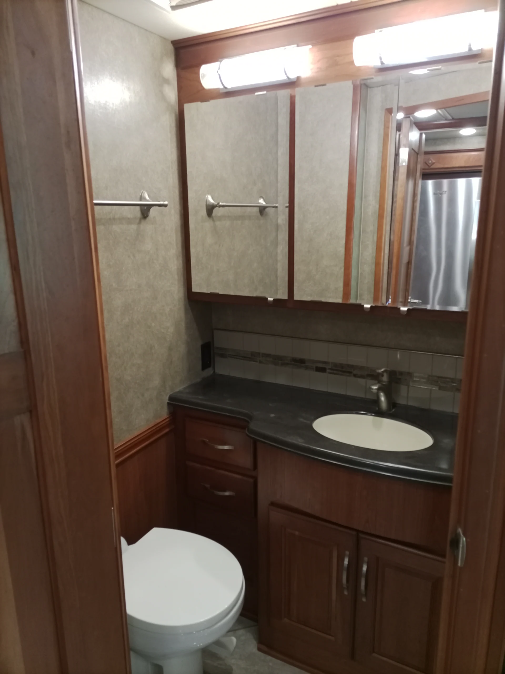 IMG_20260928_092927_068 Compact RV bathroom with wood cabinets, counter with sink, mirror, toilet, and towel rack; typical of Motorcoach and RVs.