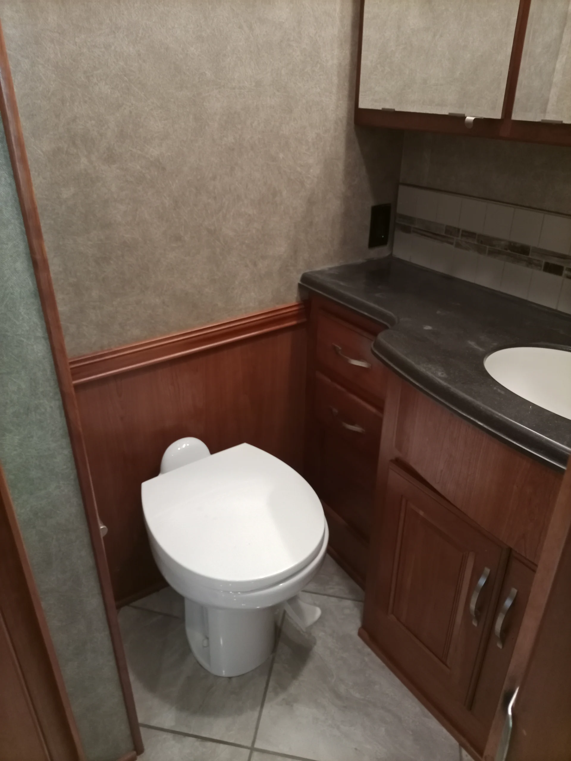 IMG_20260928_092938_756 Compact bathroom in a motorcoach featuring a toilet, dark countertop, and wood cabinetry, highlighting efficient use of space.