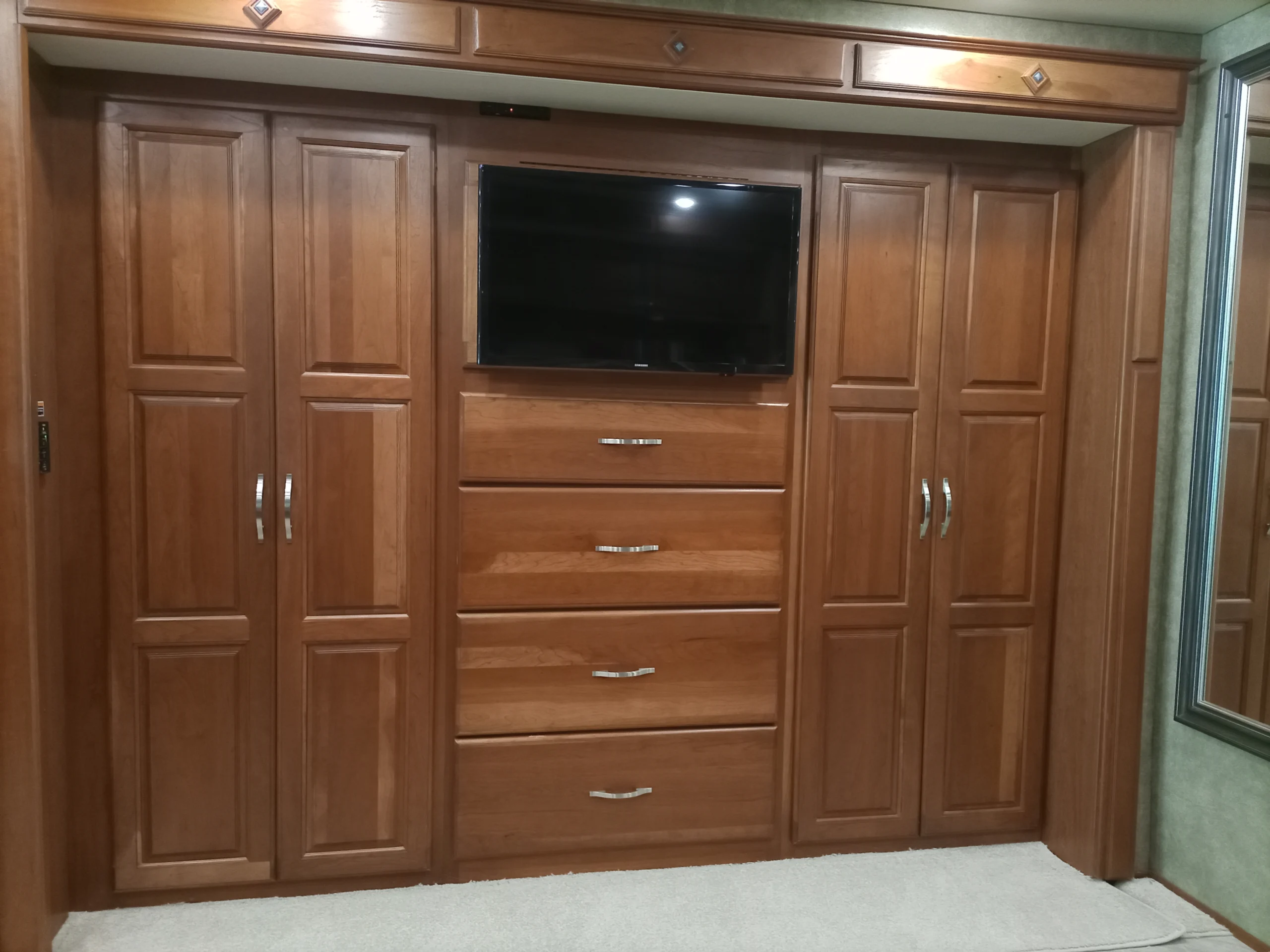 IMG_20260928_093042_310 Wooden cabinetry with TV, drawers, and closets in a motorcoach or RV interior; mirrored door to the right.