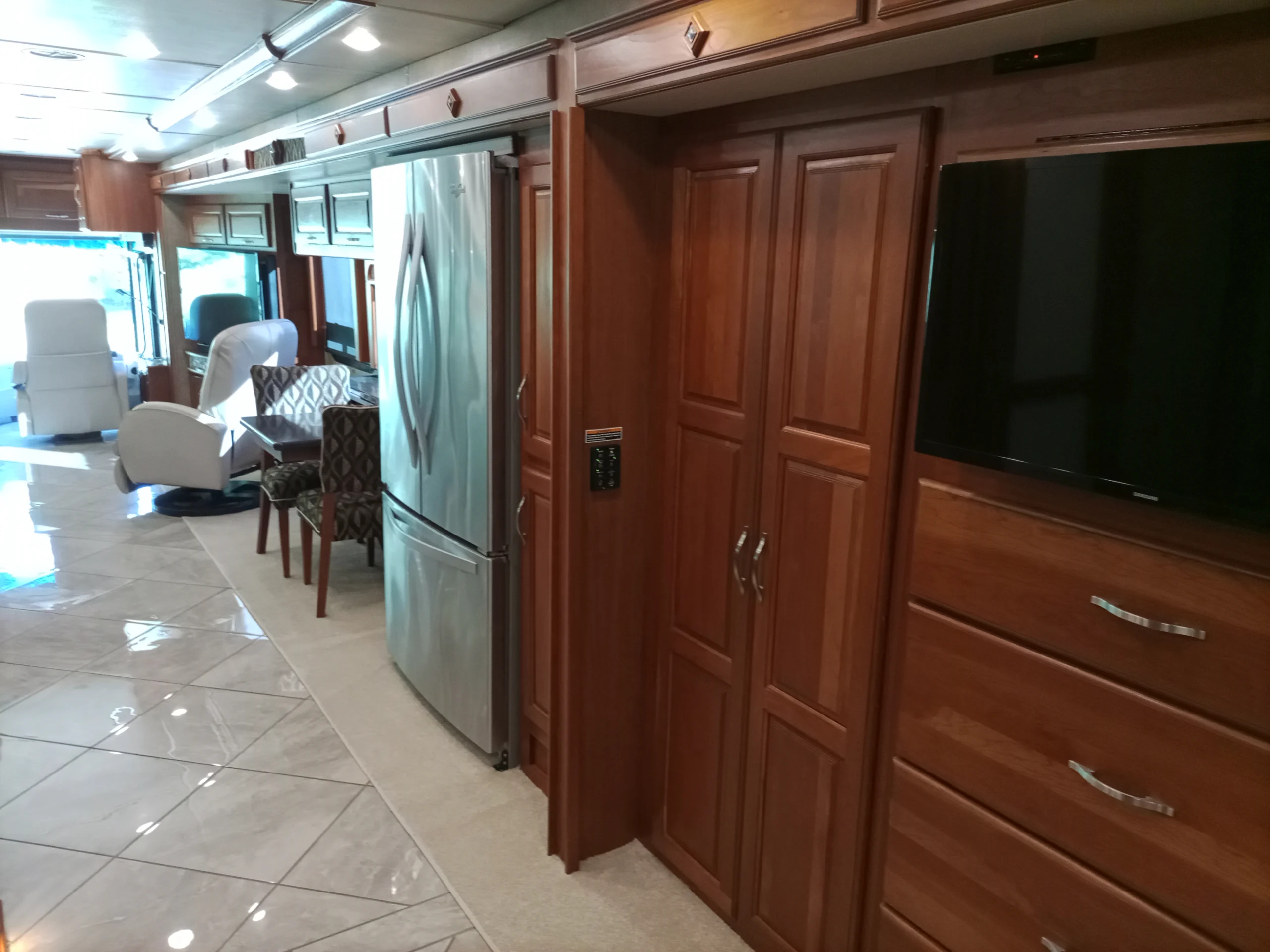 IMG_20260928_093109_508 Spacious Motorcoach interior with wood cabinets, stainless fridge, dining area, leather chairs, tile flooring, and mounted TV.