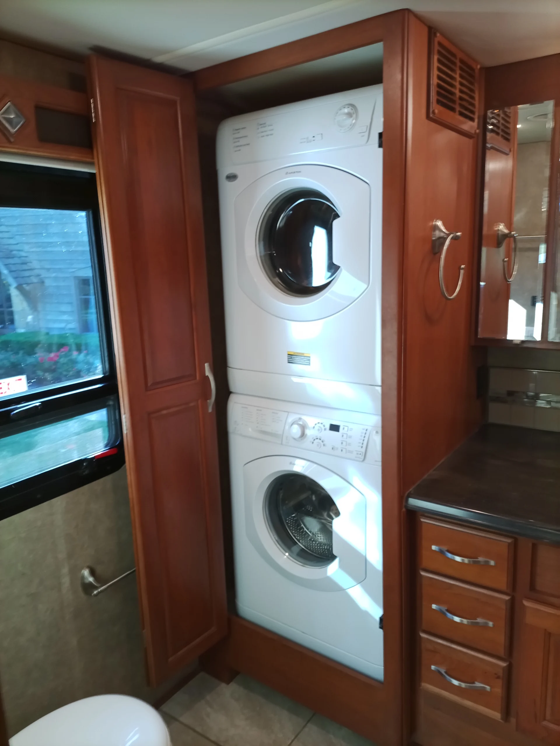 IMG_20260928_093202_979 Stacked washer and dryer in a Motorcoach and RV interior, flanked by wood cabinetry and drawers, next to a window and countertop.