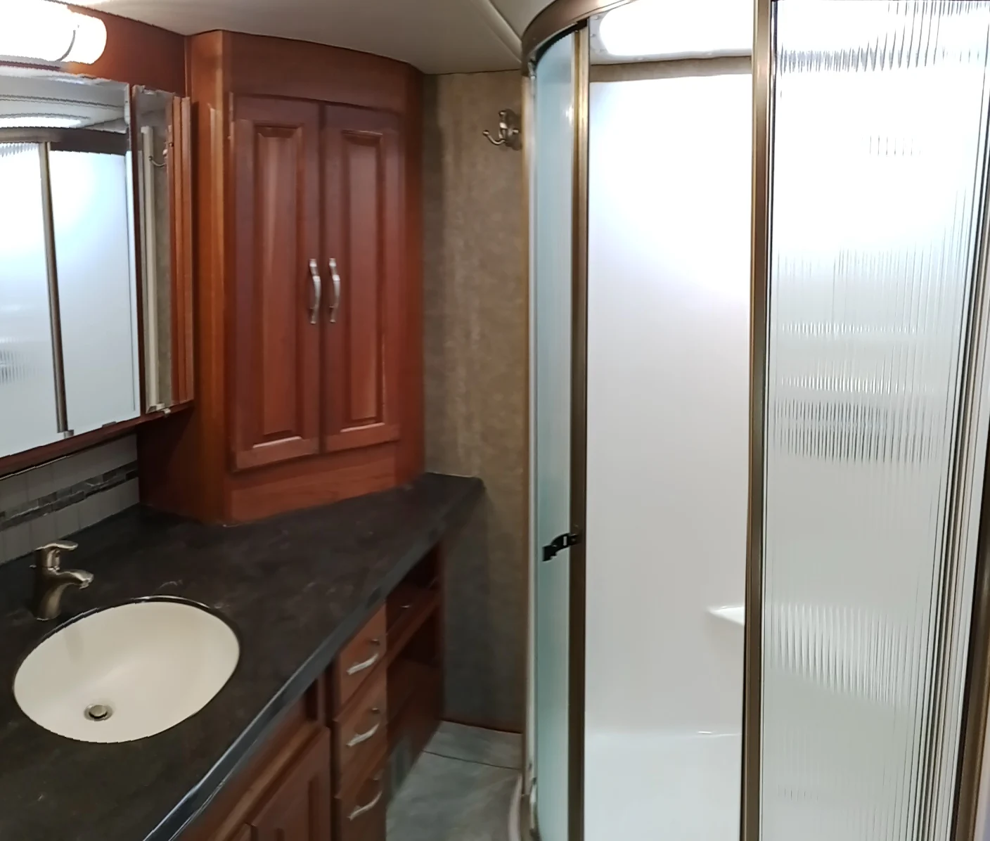 IMG_20260928_093309_1 RV bathroom with wooden cabinetry, dark countertop, under-mount sink, mirrored cabinet, and a frosted glass shower enclosure.