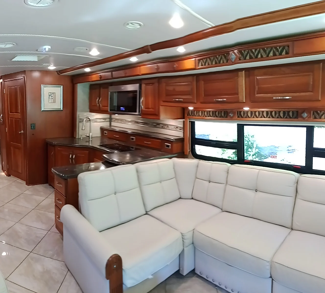 IMG_20260928_093609_6 Spacious Motorcoach interior with a white sectional sofa, wooden cabinets, sleek kitchen area, and large windows providing natural light.