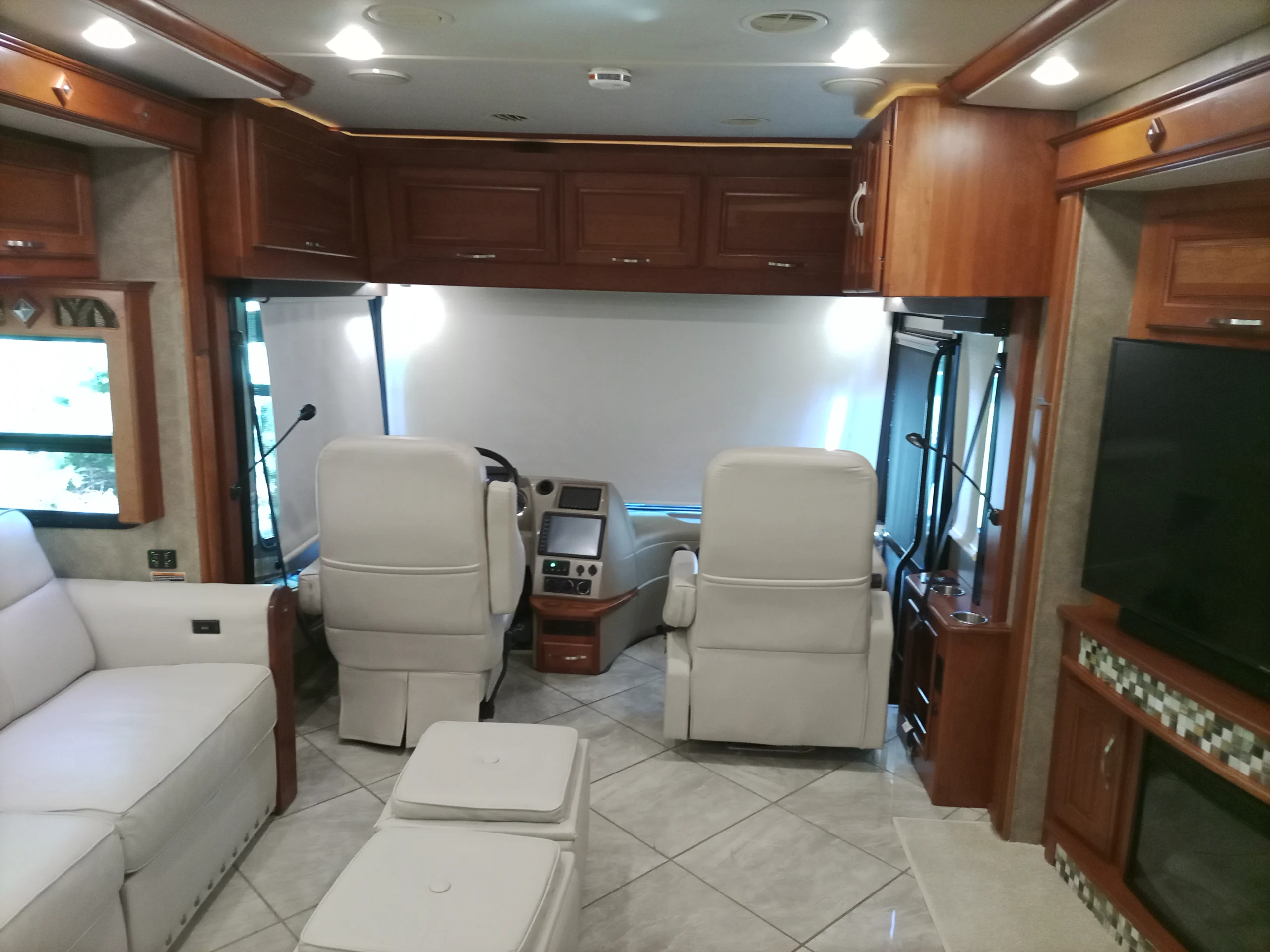 IMG_20260928_093911_530 Luxury motorcoach interior featuring cream leather seating, wooden cabinetry, a large TV, and spacious living area.