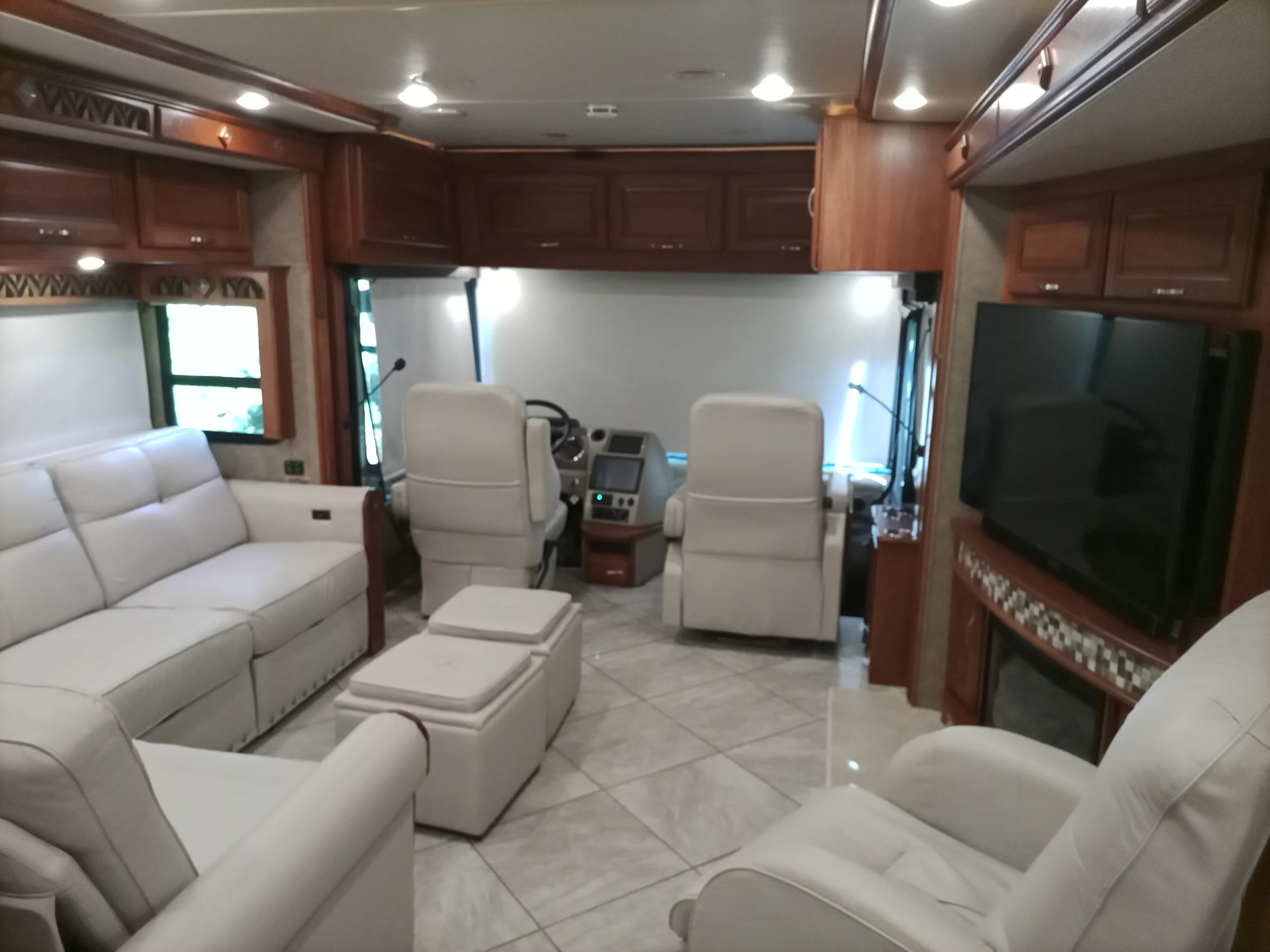 IMG_20260928_094115_290 Luxurious Motorcoach interior featuring plush white seating, wood cabinets, tiled flooring, large TV, and driving area with a panoramic windshield.