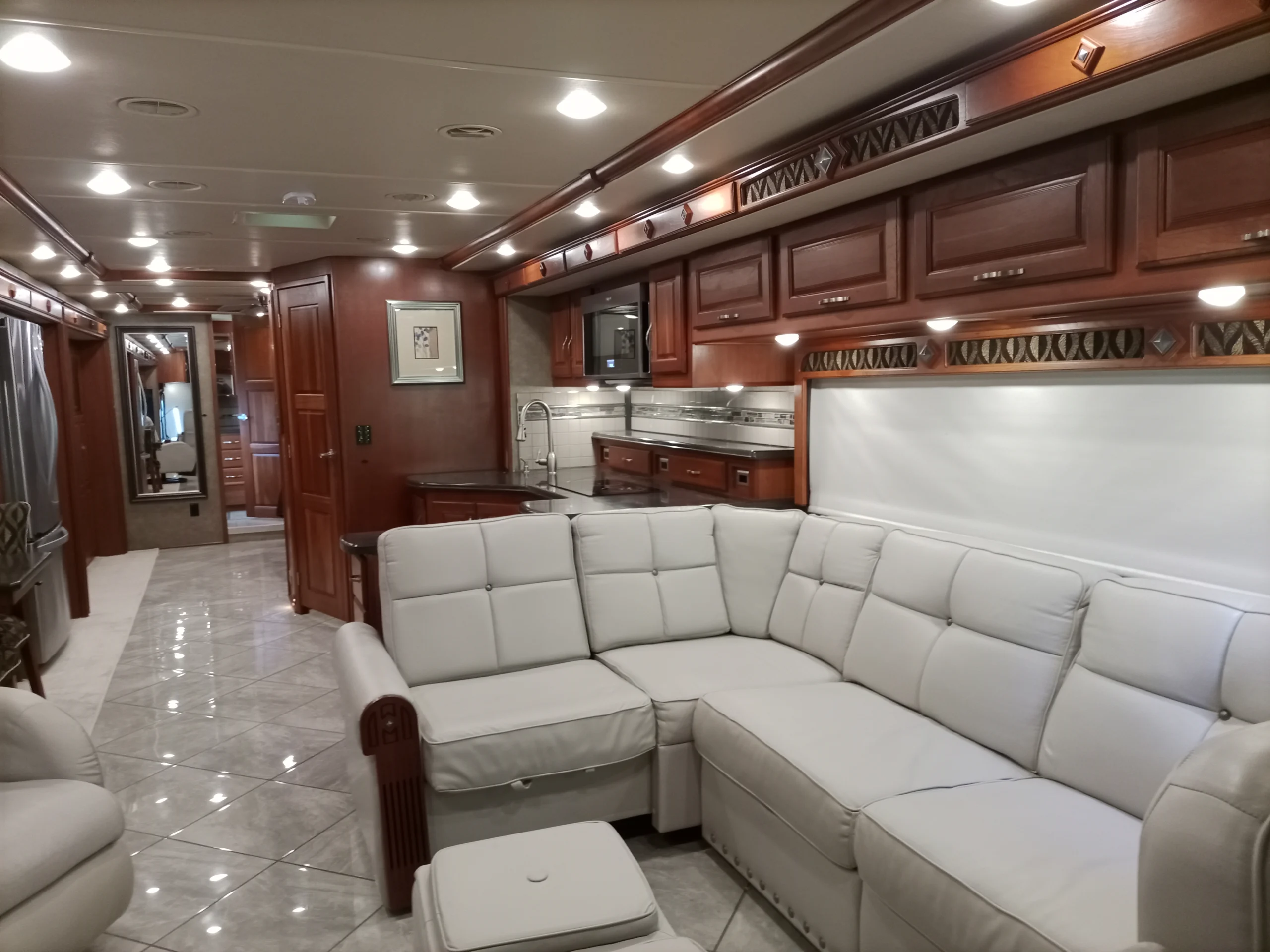 IMG_20260928_094223_341 Luxurious motorcoach interior with white leather sectional, wooden cabinets, stainless steel kitchen, and glossy tiled floor.