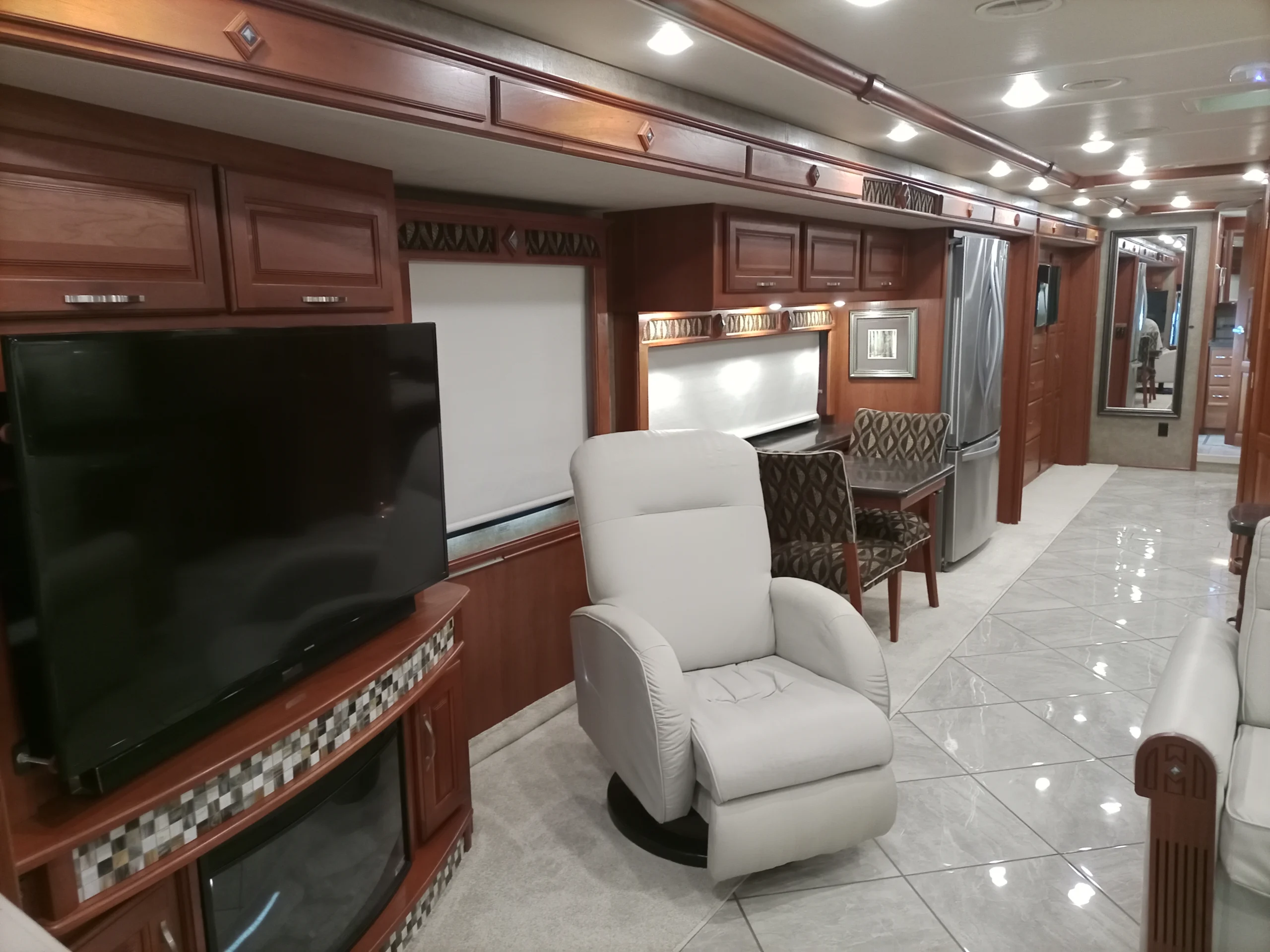 IMG_20260928_094230_028 Luxurious motorcoach interior featuring a plush recliner, large TV, elegant wood cabinetry, and spacious kitchen with stainless steel appliances.