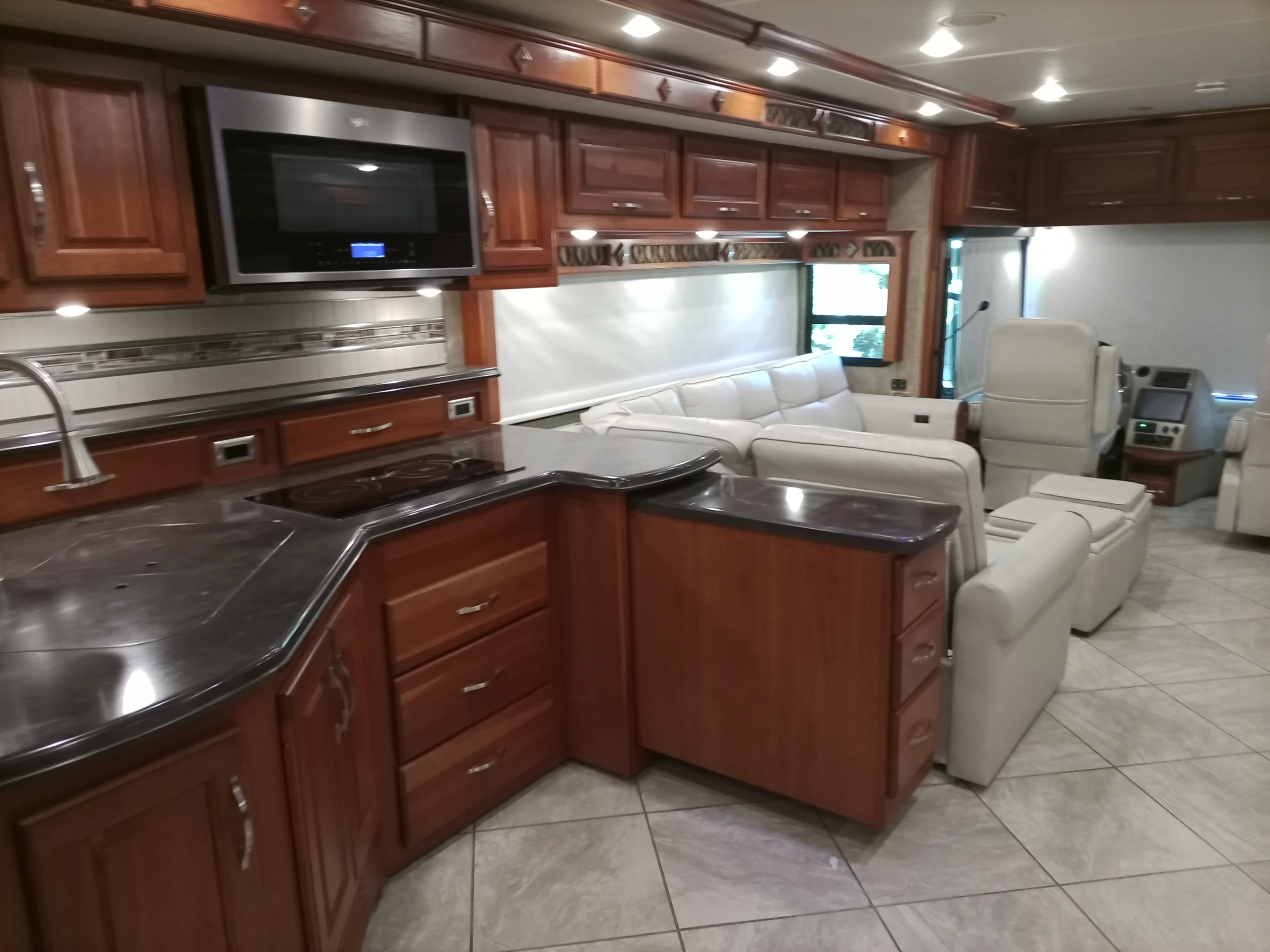 IMG_20260928_094254_453 Luxurious Motorcoach interior with wooden cabinets, plush white seating, modern kitchenette, and tiled floors, creating a spacious, elegant look.