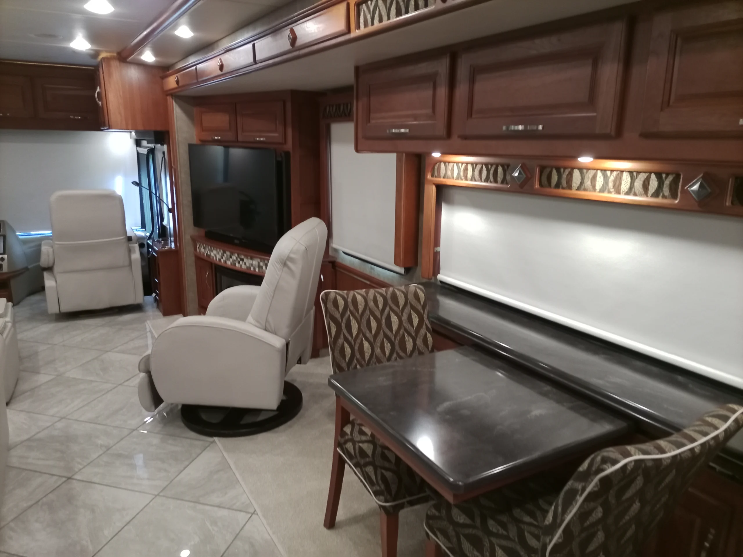 IMG_20260928_094259_710 Luxurious Motorcoach and RVs interior with leather chairs, dining table, flat-screen TV, and wooden cabinets.