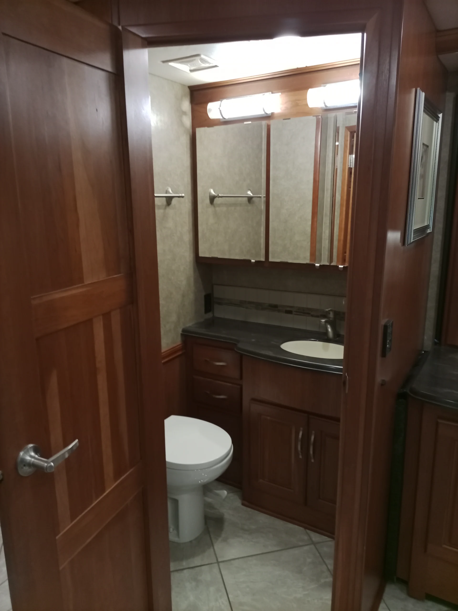 IMG_20260928_094322_576 Wood-paneled RV bathroom with vanity, mirror cabinet, sink, and toilet, showcasing Motorcoach aesthetics.