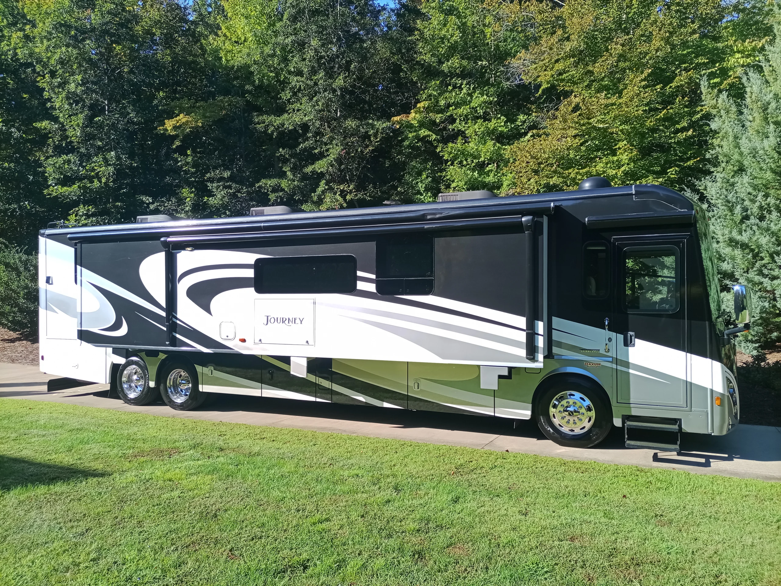 IMG_20260928_094842_504 Black and white motorcoach parked on a driveway, surrounded by lush green trees and grass.