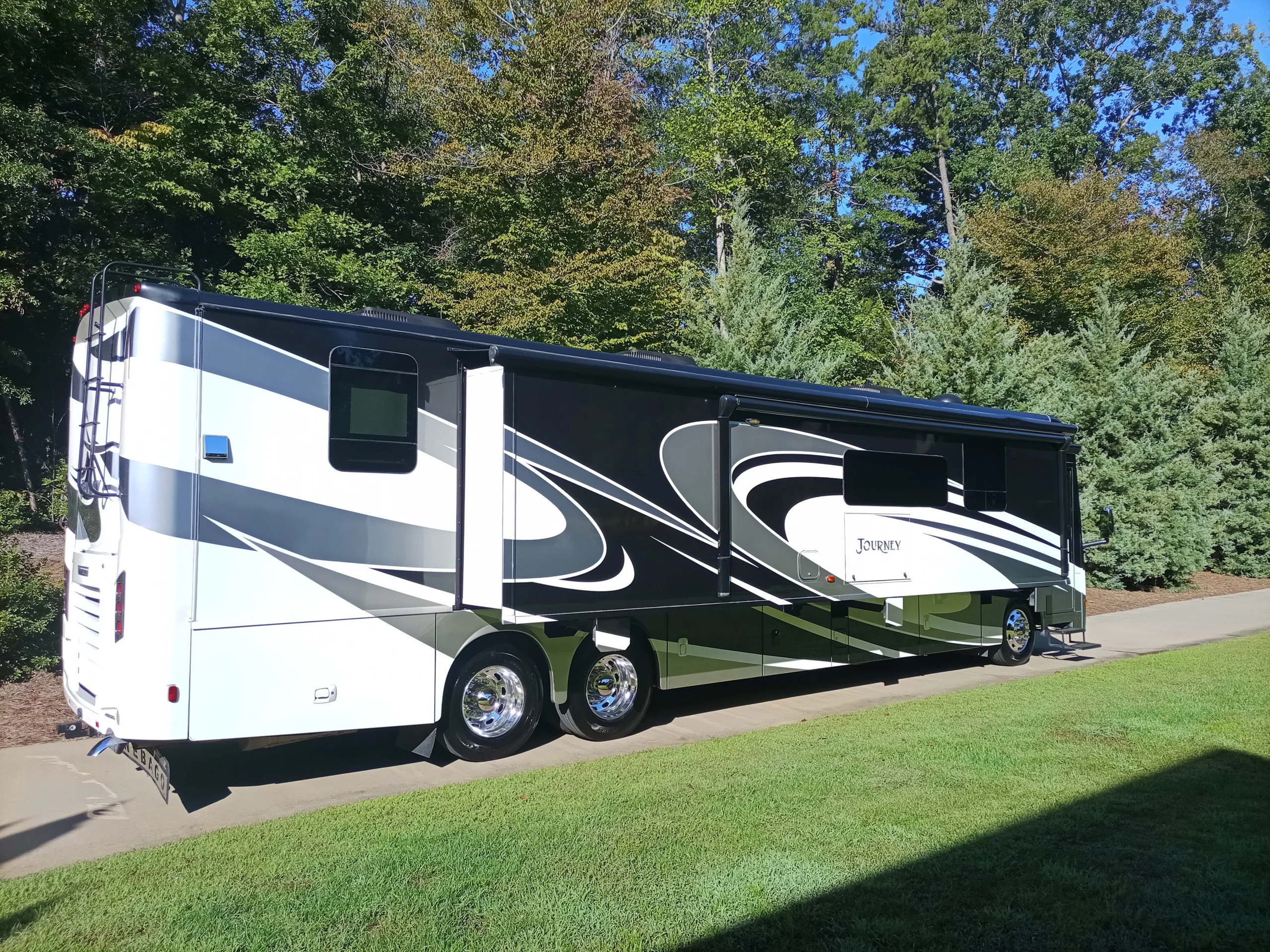 IMG_20260928_094913_572 Luxury motorcoach parked on a driveway, surrounded by lush green trees and grass, featuring sleek white and black exterior design.